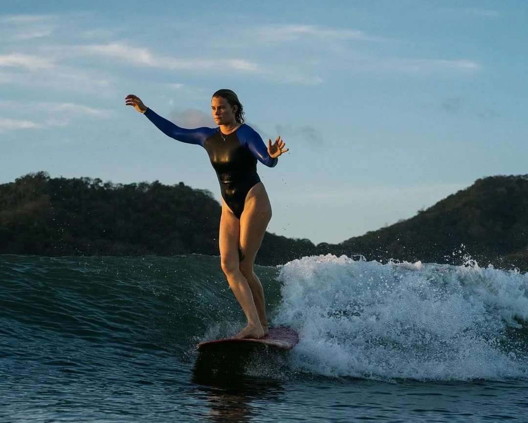 Woman surfing on a wave during sunset with a background of hills and a cloudy sky.