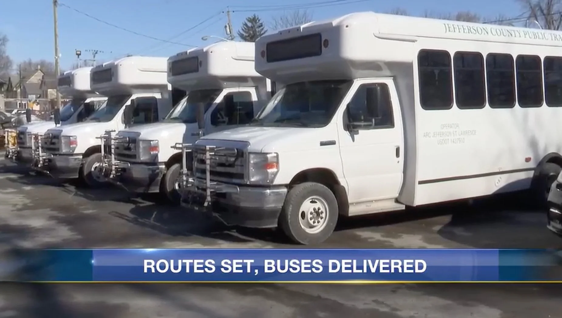 Multiple white buses parked in a lot, with the front bus in the foreground showing a sign that reads "Jefferson County Public Tr"); a news banner at the bottom says "ROUTES SET, BUSES DELIVERED."