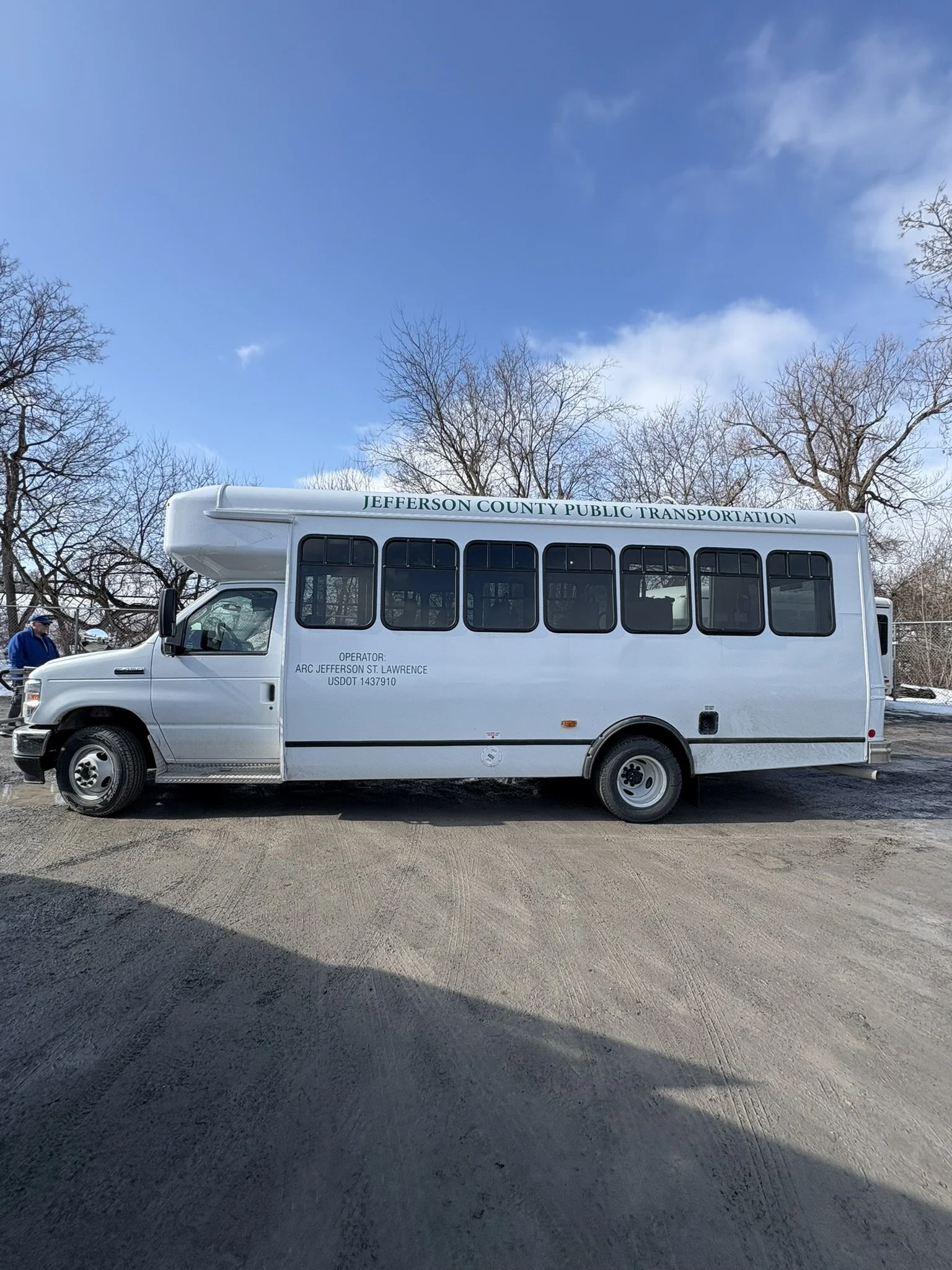 A white Jefferson County Public Transportation shuttle bus parked on a gravel ground with leafless trees in the background and a partly cloudy blue sky.