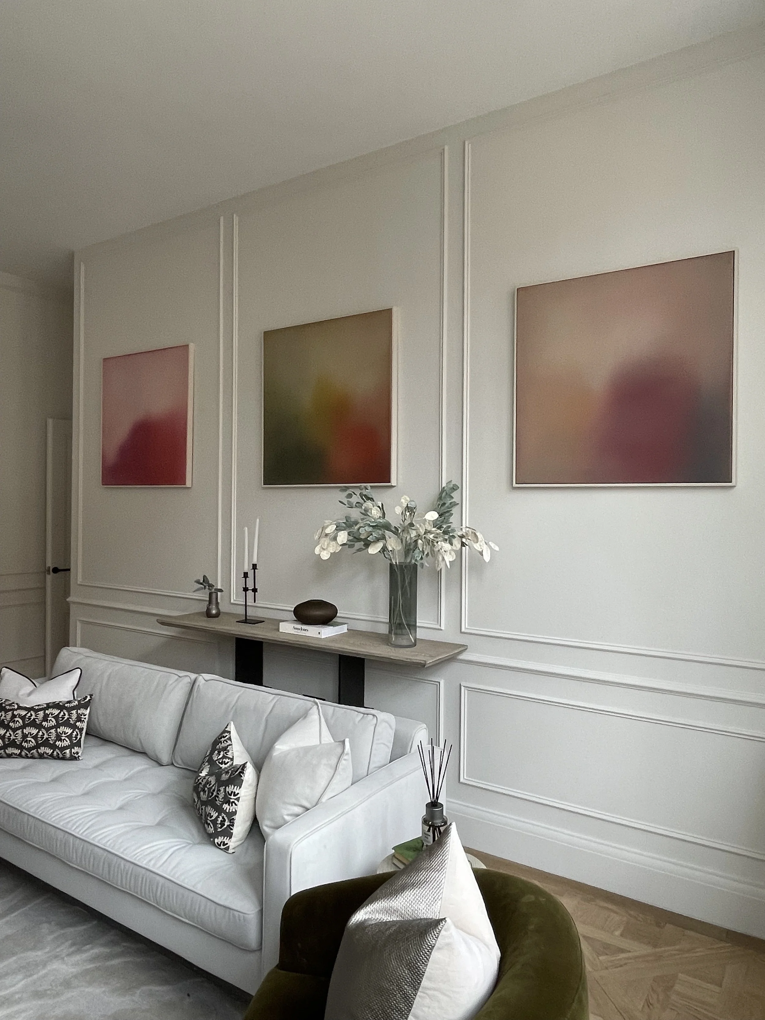 Living room with white sofa, decorative pillows, a round velvet armchair, a console table with vases and candles, a large vase with white flowers, and three abstract framed artworks on the wall.