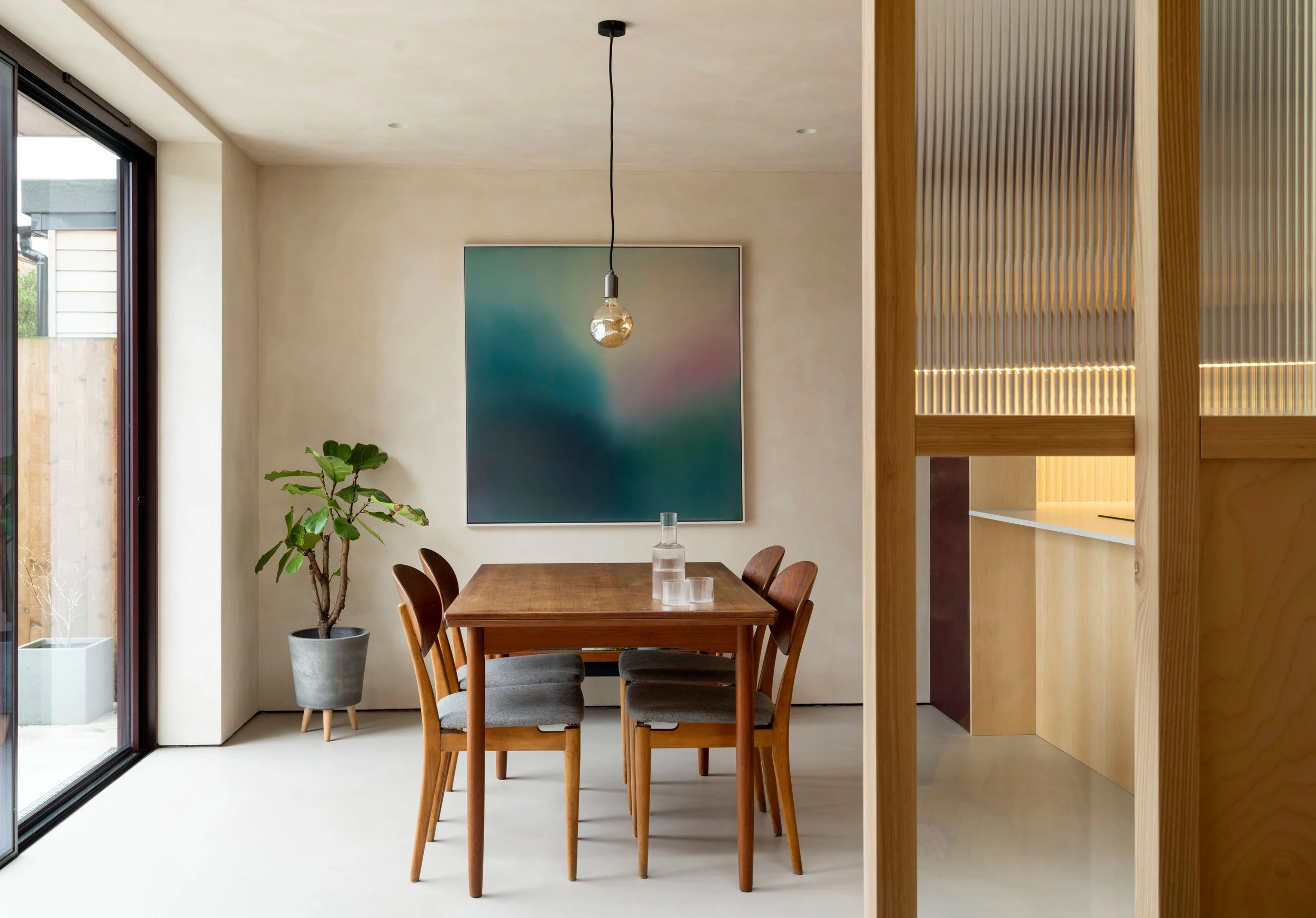 Modern dining area with a wooden table, six chairs, a potted plant, a glass bottle, a framed abstract art piece on the wall, and a hanging light bulb. Large sliding glass doors on the left lead outside, with natural light illuminating the room.