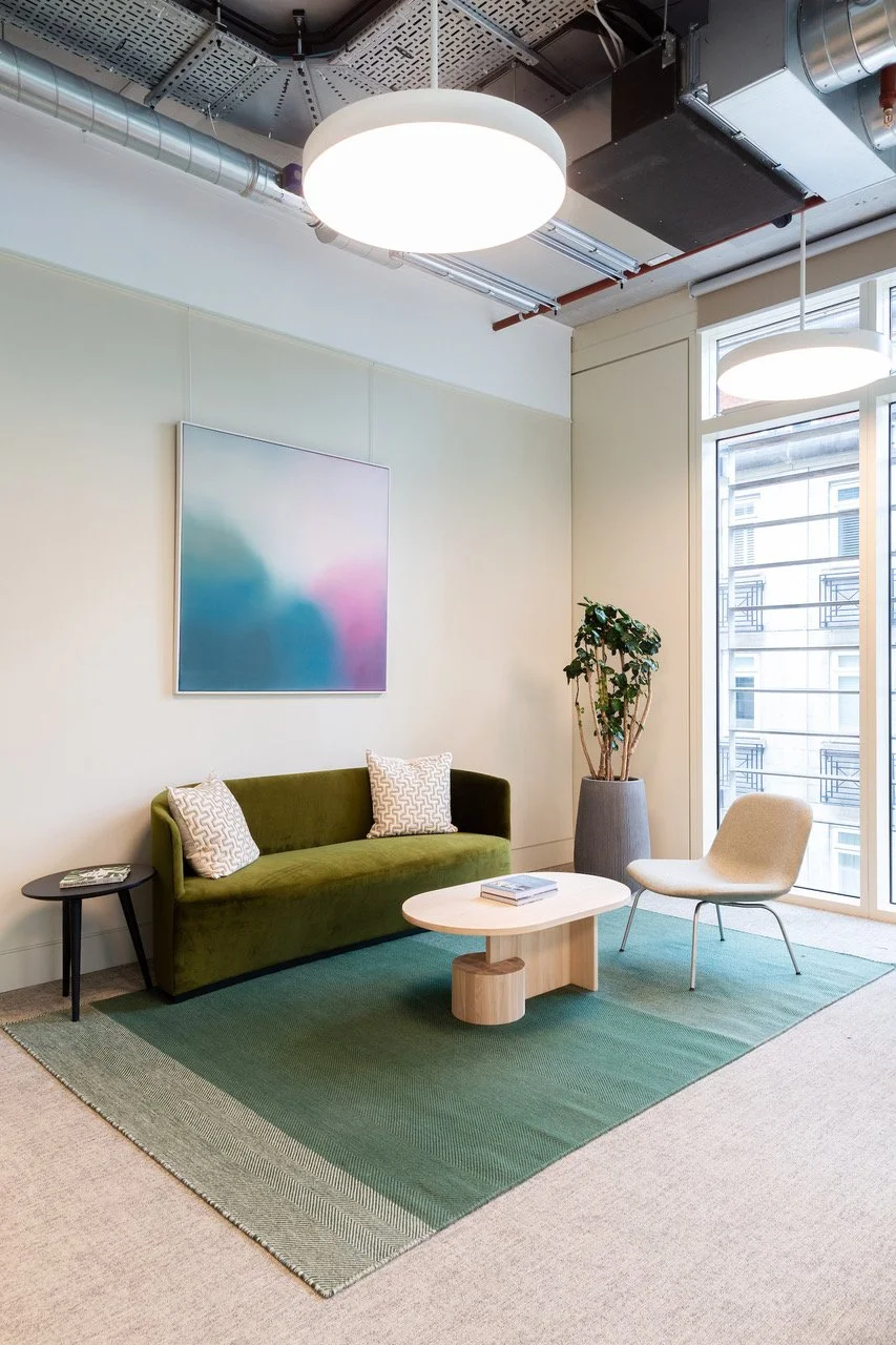 Modern lounge area with green velvet sofa, white armchair, small wooden coffee table, black side table, potted plant, abstract artwork on the wall, and large windows letting in natural light.
