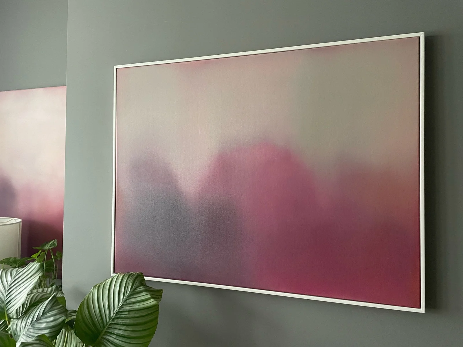 A large, square, abstract art piece with a pink gradient, framed and hanging on a gray wall in a room with green plants.