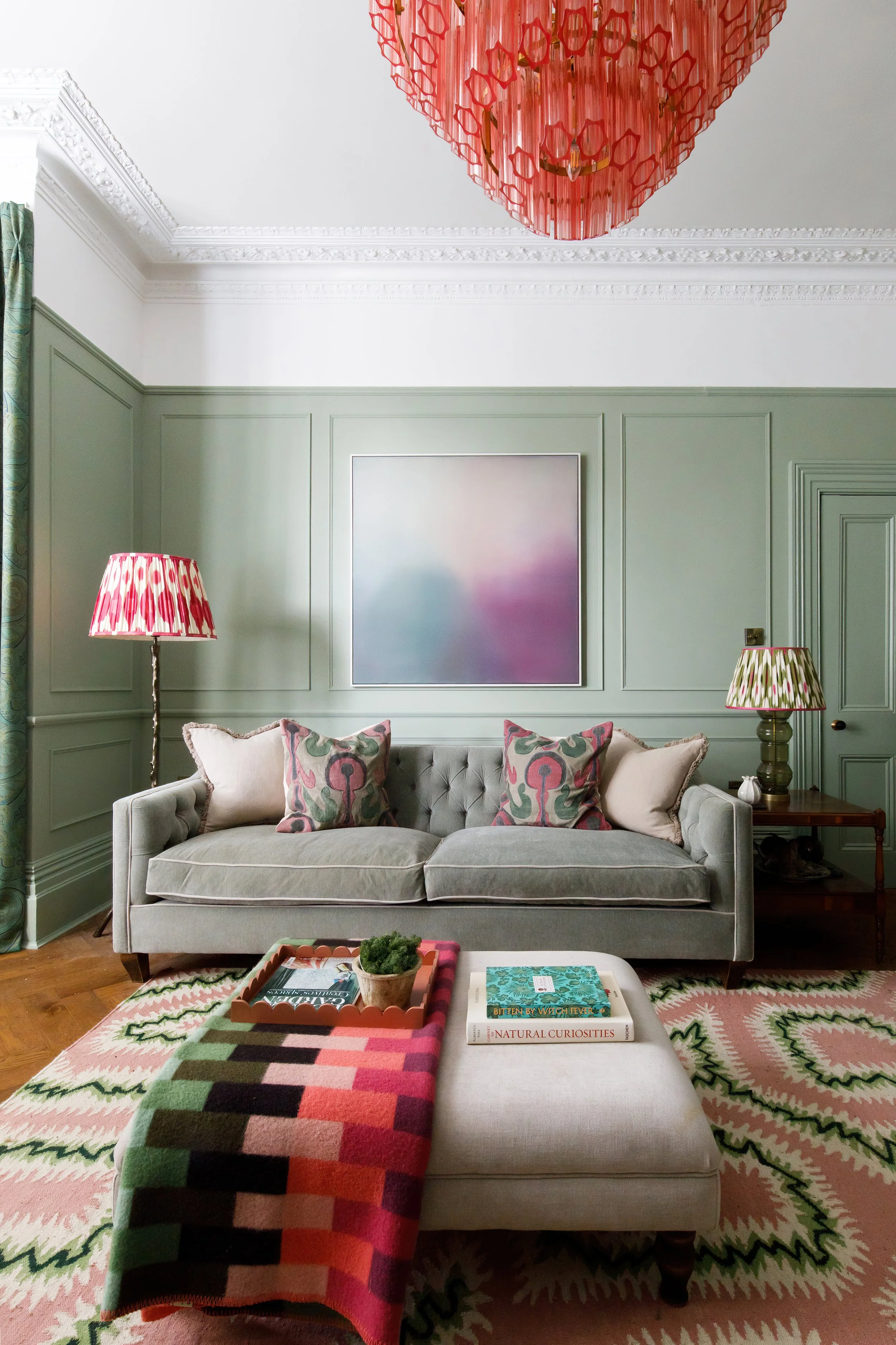 Living room with a grey couch decorated with patterned pillows, a colorful geometric throw blanket, a wooden side table with lamps, a pink and green patterned rug, a coffee table with books, a pink chandelier, and green wall paneling.