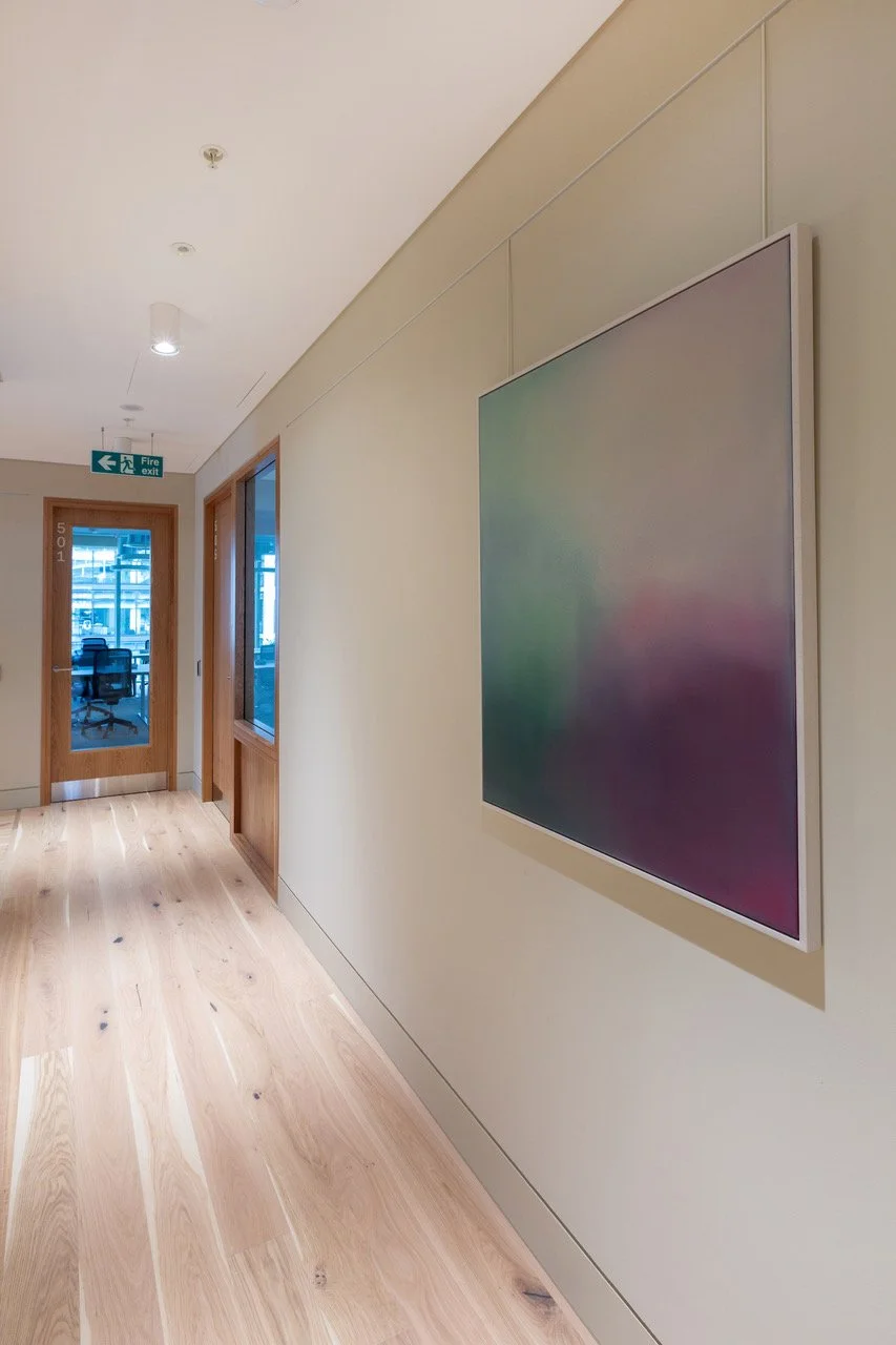 Empty hallway with light wood floor, cream walls, a colorful framed art piece, and a glass door leading to another room with a view of outside buildings.