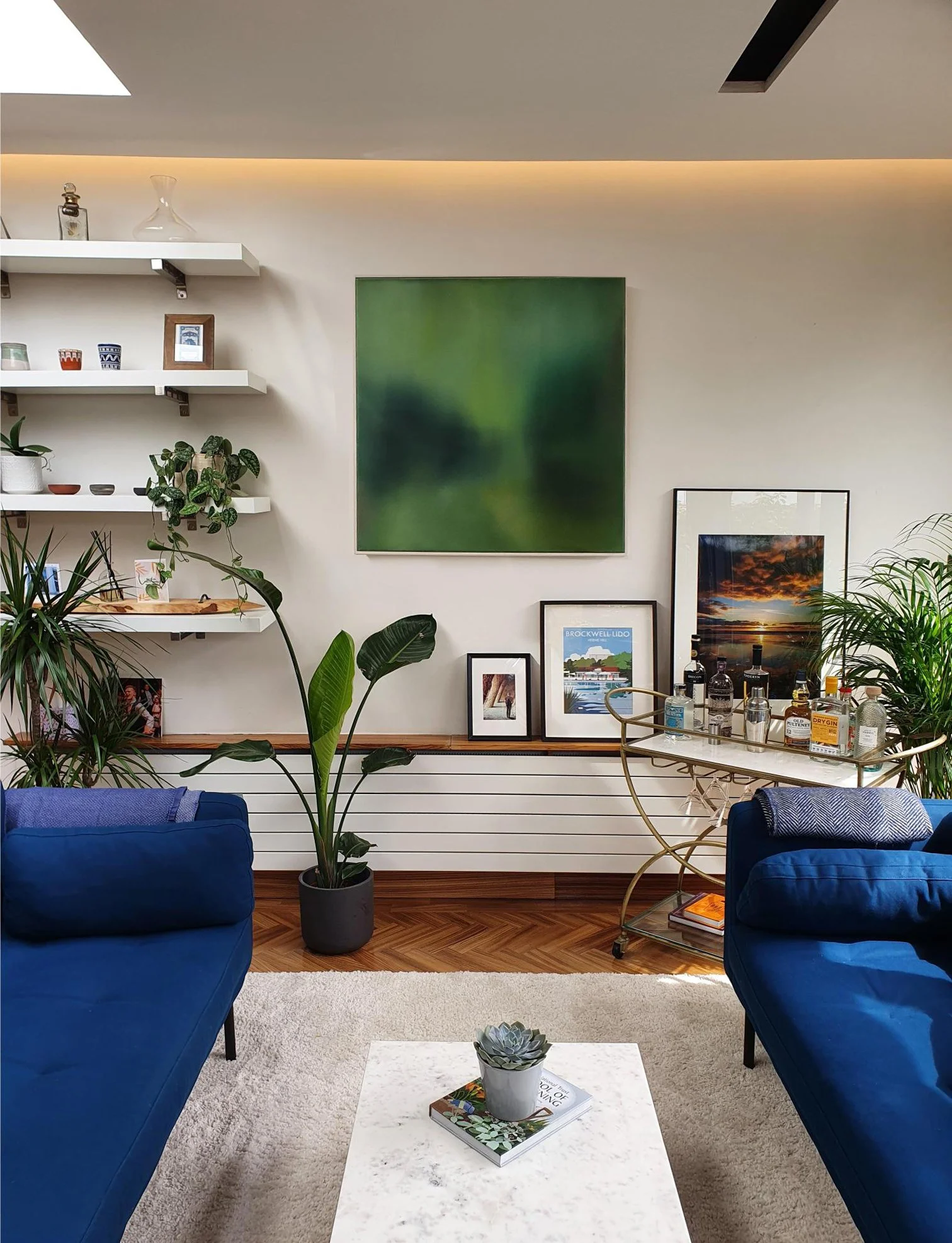 Living room with white walls, blue sofas, and wooden flooring. Decor includes framed pictures, a green potted plant, and a white coffee table with a plant and books. Artwork on the wall includes an abstract green painting and framed sunset photos.