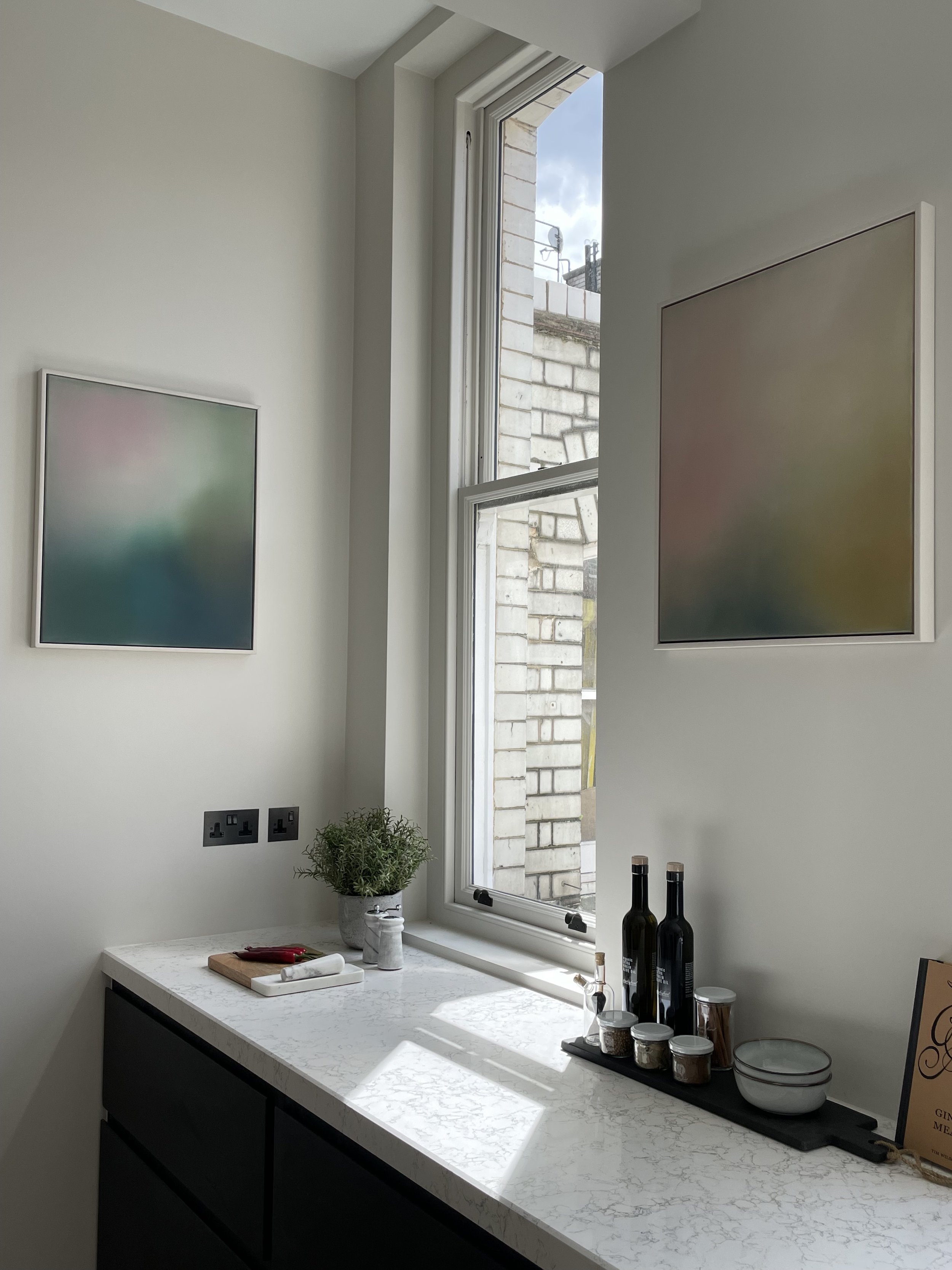 A kitchen countertop with a potted plant, a cutting board, soap dispenser, and three spice jars. Above the counter is a window with sunlight casting shadows, and two framed pictures on the light-colored wall.