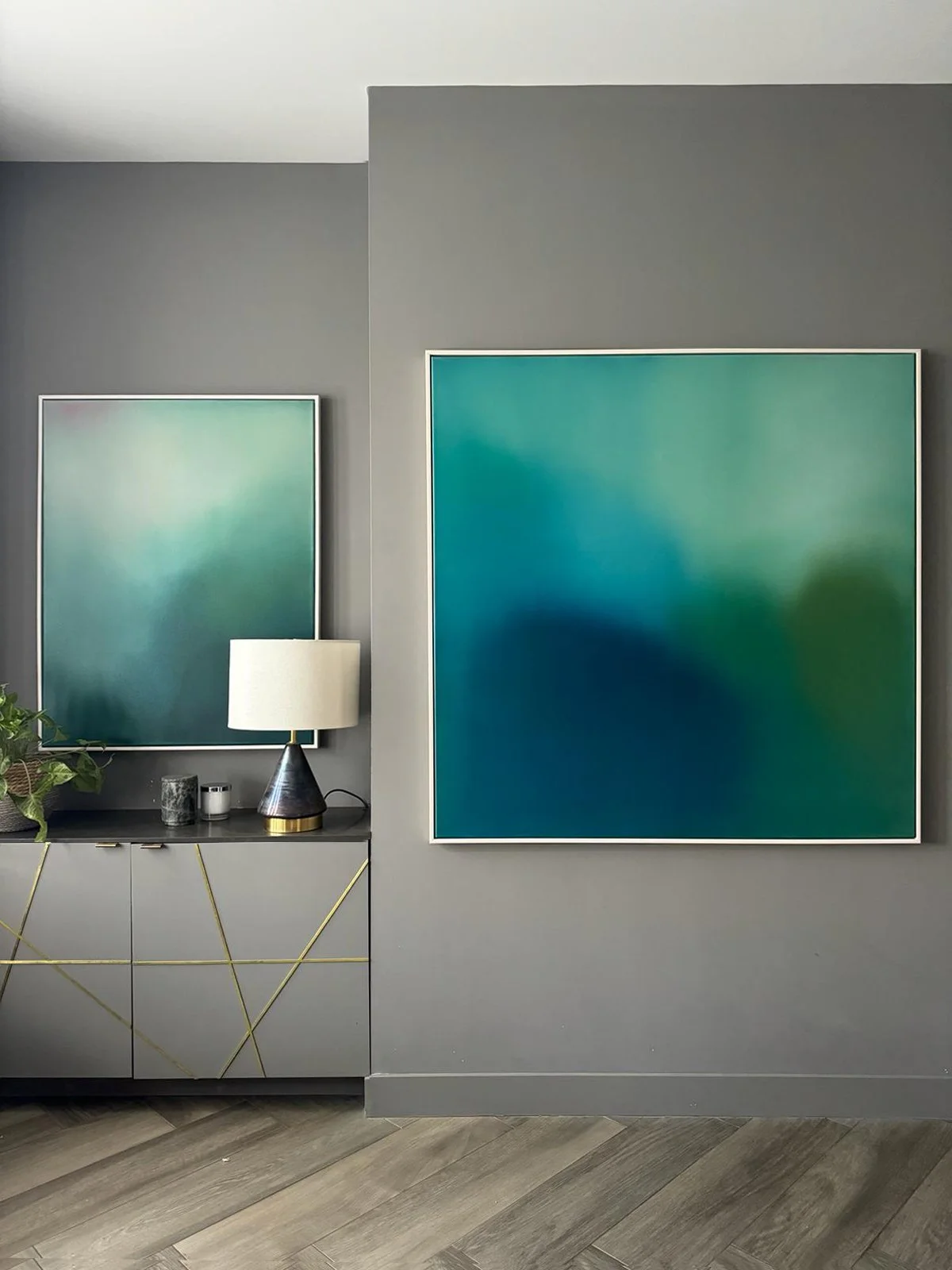 Living room with gray walls, two large abstract teal and green artwork, a gray cabinet with a gold geometric pattern, a black and gold table lamp, a potted plant, and some small decorative items, with wood plank flooring.