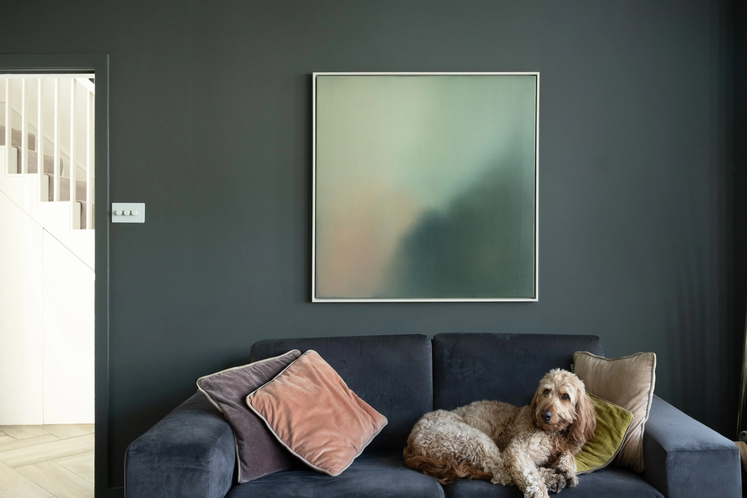 A living room with grey walls, a framed oil painting, a dark gray sofa with pillows, and a dog lying on the sofa.