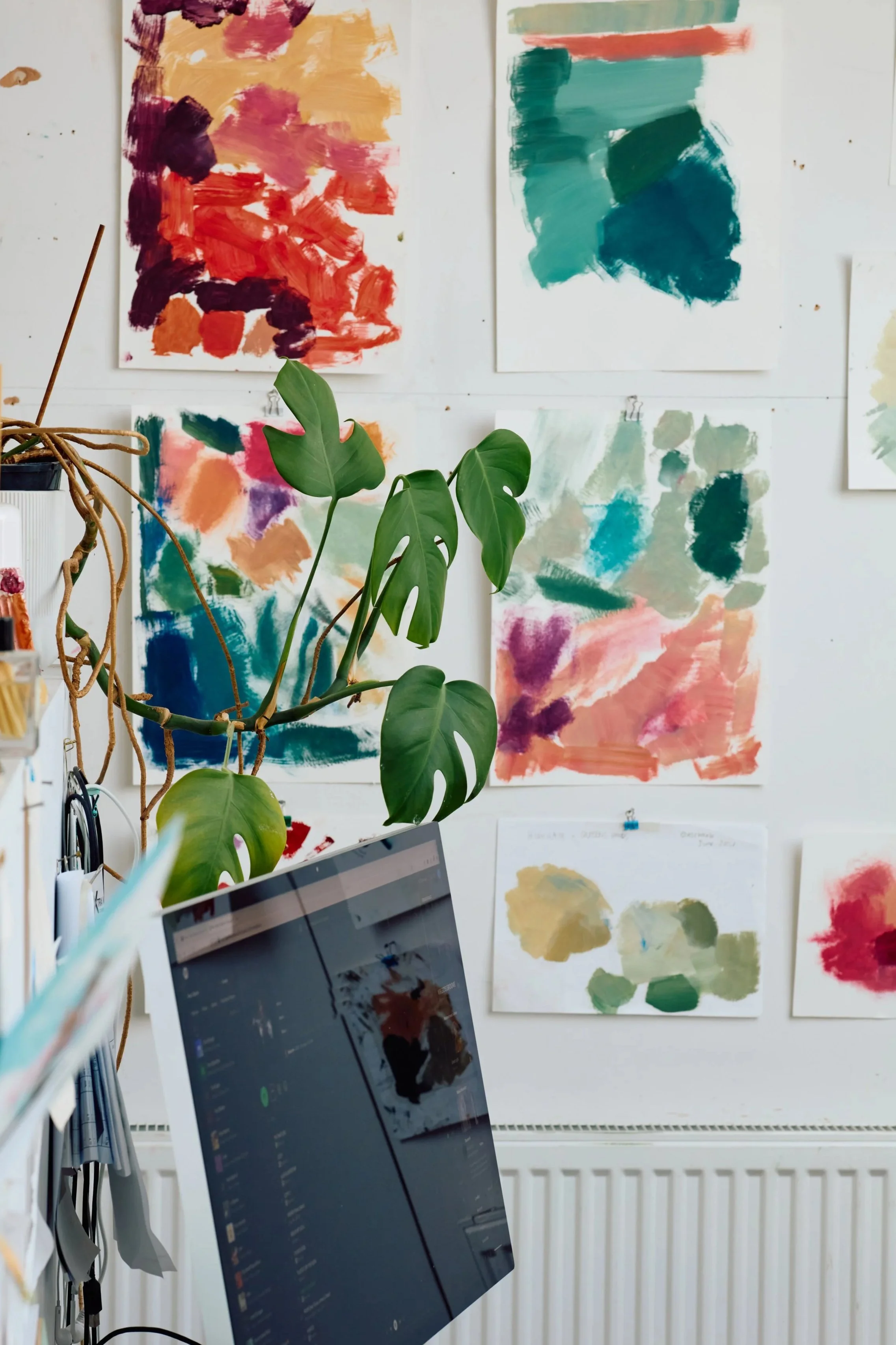 Wall decorated with multiple abstract watercolor paintings in various colors such as red, orange, green, blue, and purple, with a potted plant in the foreground and a computer screen partially visible.