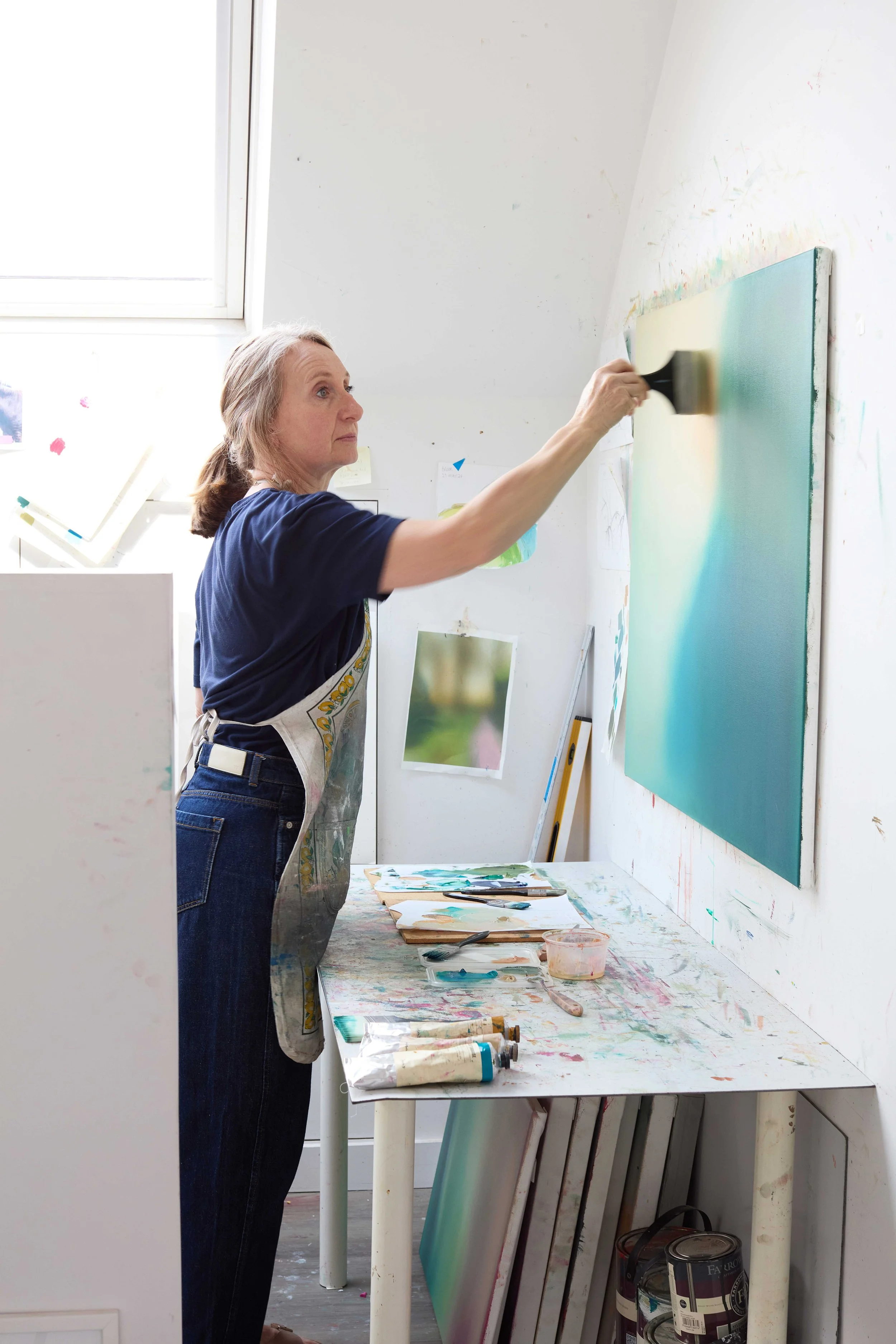 A woman painting on a large canvas in an art studio with paint supplies on a table.