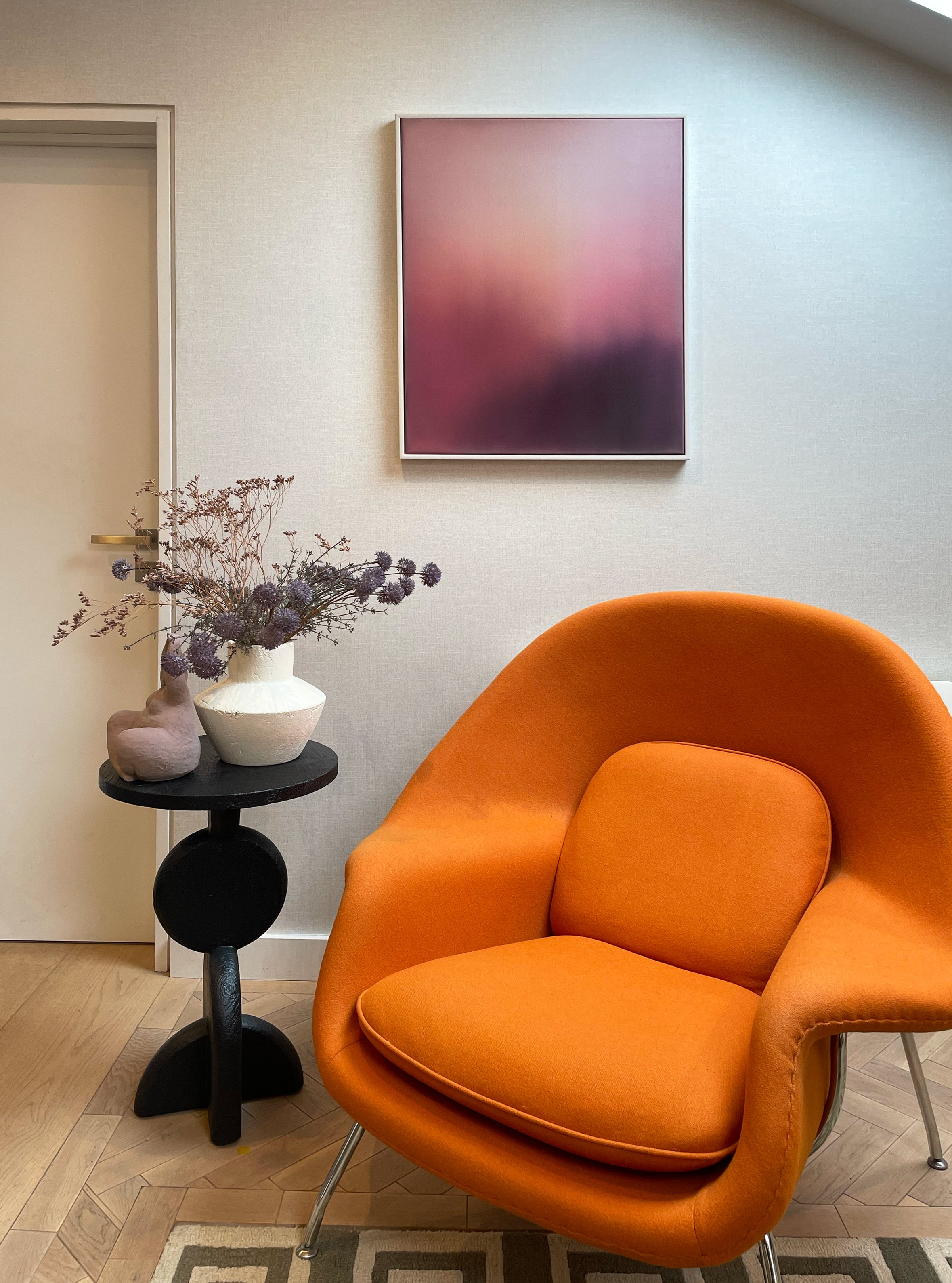 A cozy orange armchair with metal legs in a room with beige walls, next to a black side table holding a white vase with dried purple flowers. A modern abstract pink and purple painting hangs on the wall, and a doorway is visible on the left. The floor is light wood with a patterned area rug.