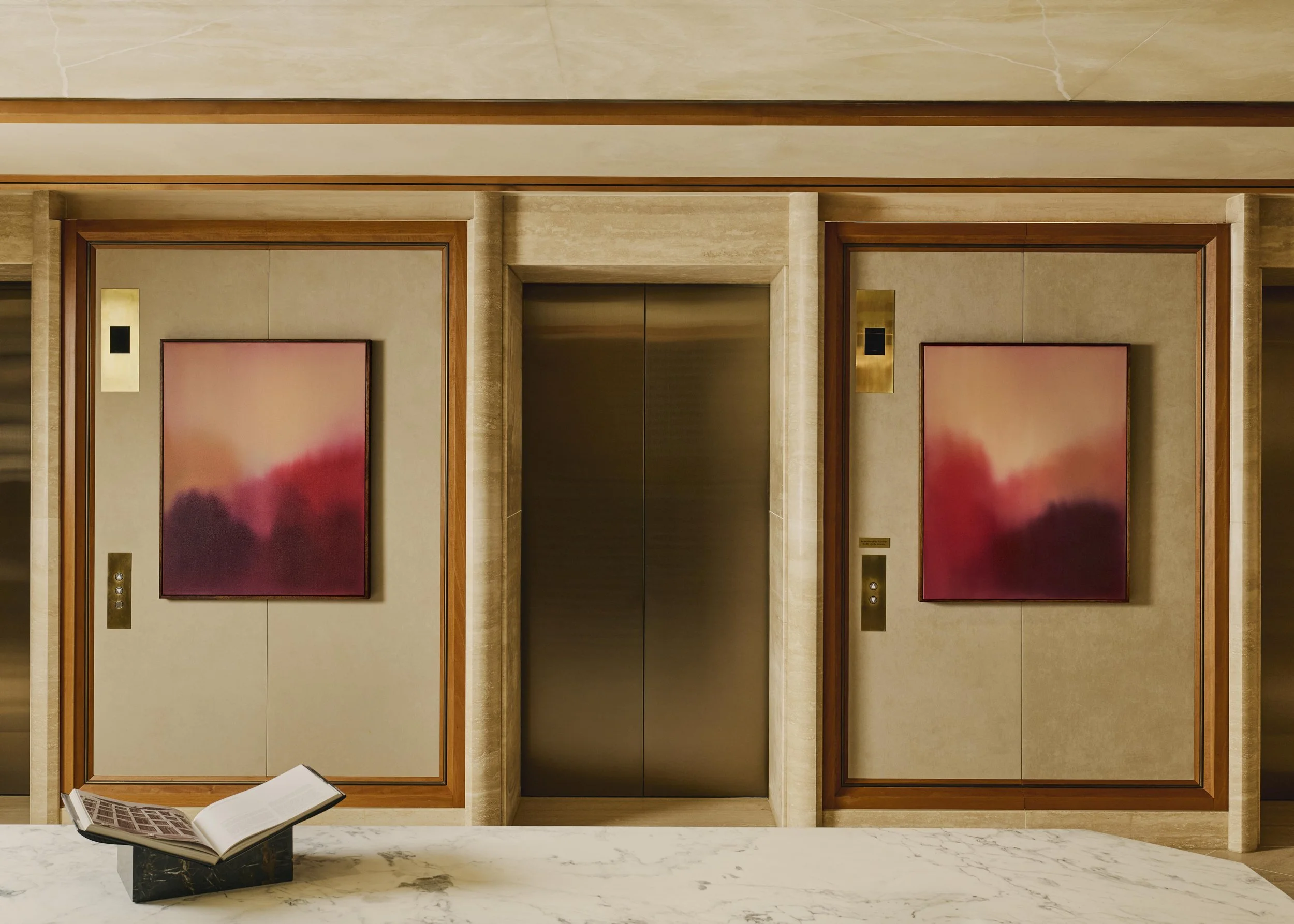 Two closed elevator doors with wood trim, beige walls, and gold elevator call buttons. A marble countertop with an open book on a stand in the foreground.