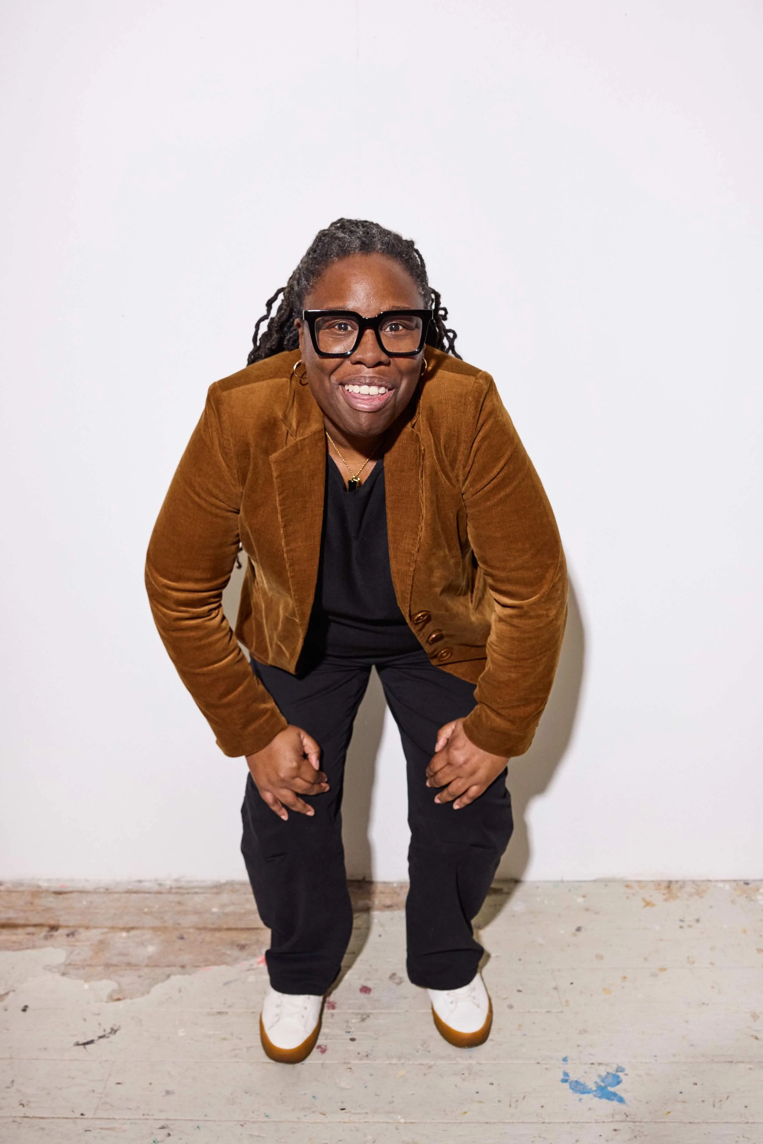 A smiling woman with locks and glasses, wearing a brown jacket, black shirt, dark pants, and white sneakers, leaning forward with hands clenched in front of a plain white wall.