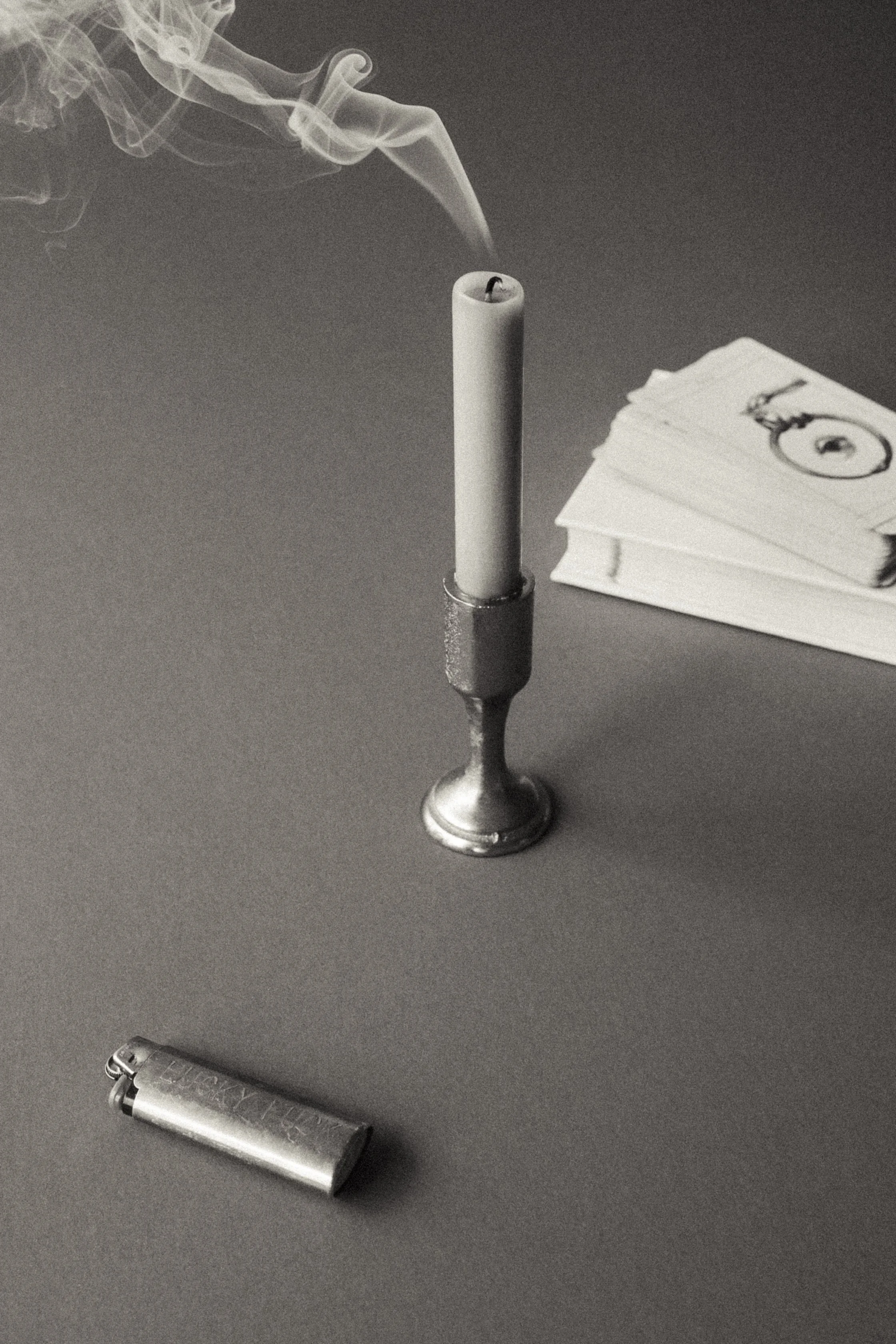 A black and white photo of a lit cigarette and a lighter on a gray surface, with a candle and a deck of cards in the background.