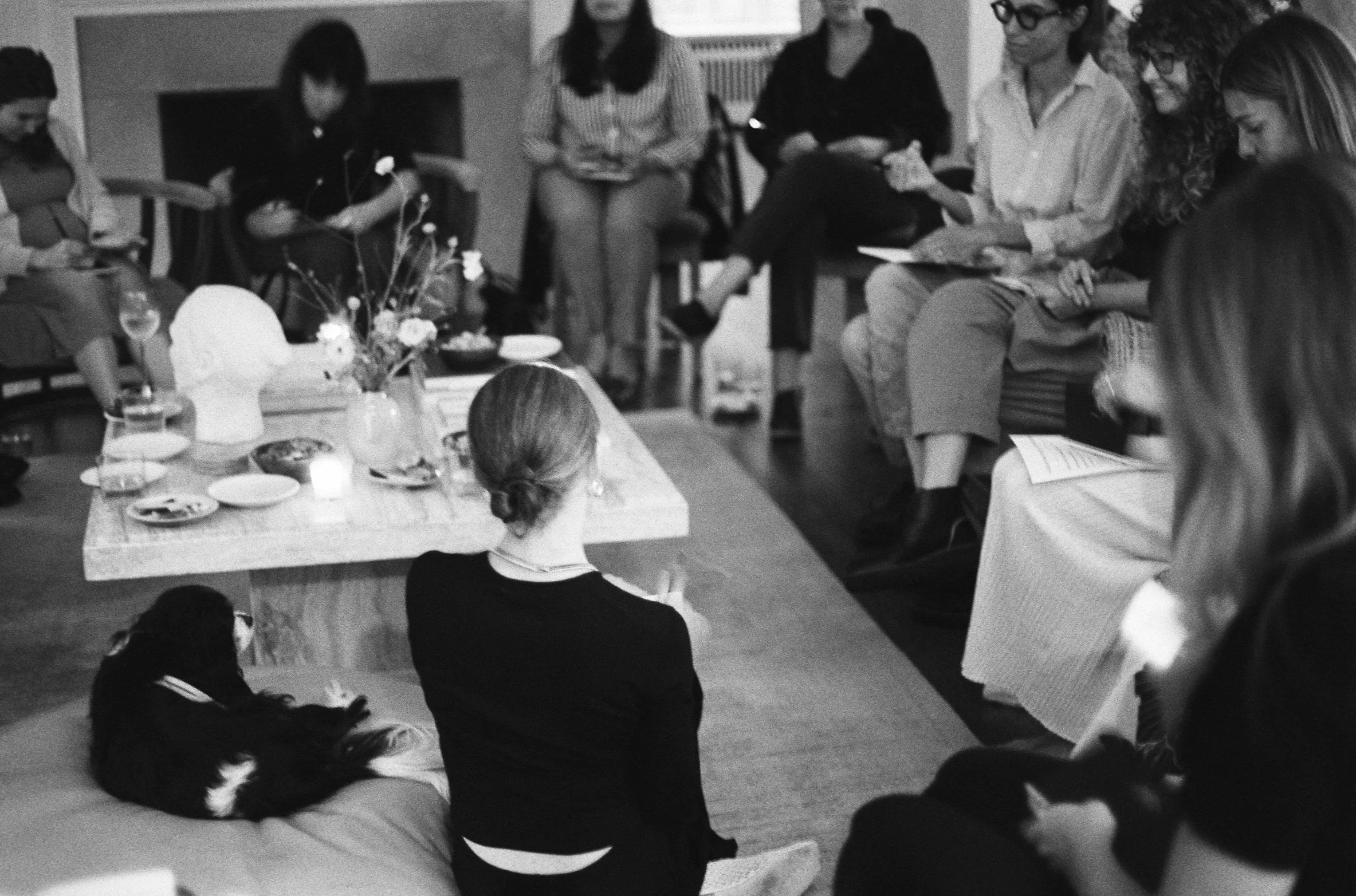 Group of women gathered around a table, some sitting and some standing, engaged in conversation, in a social setting.