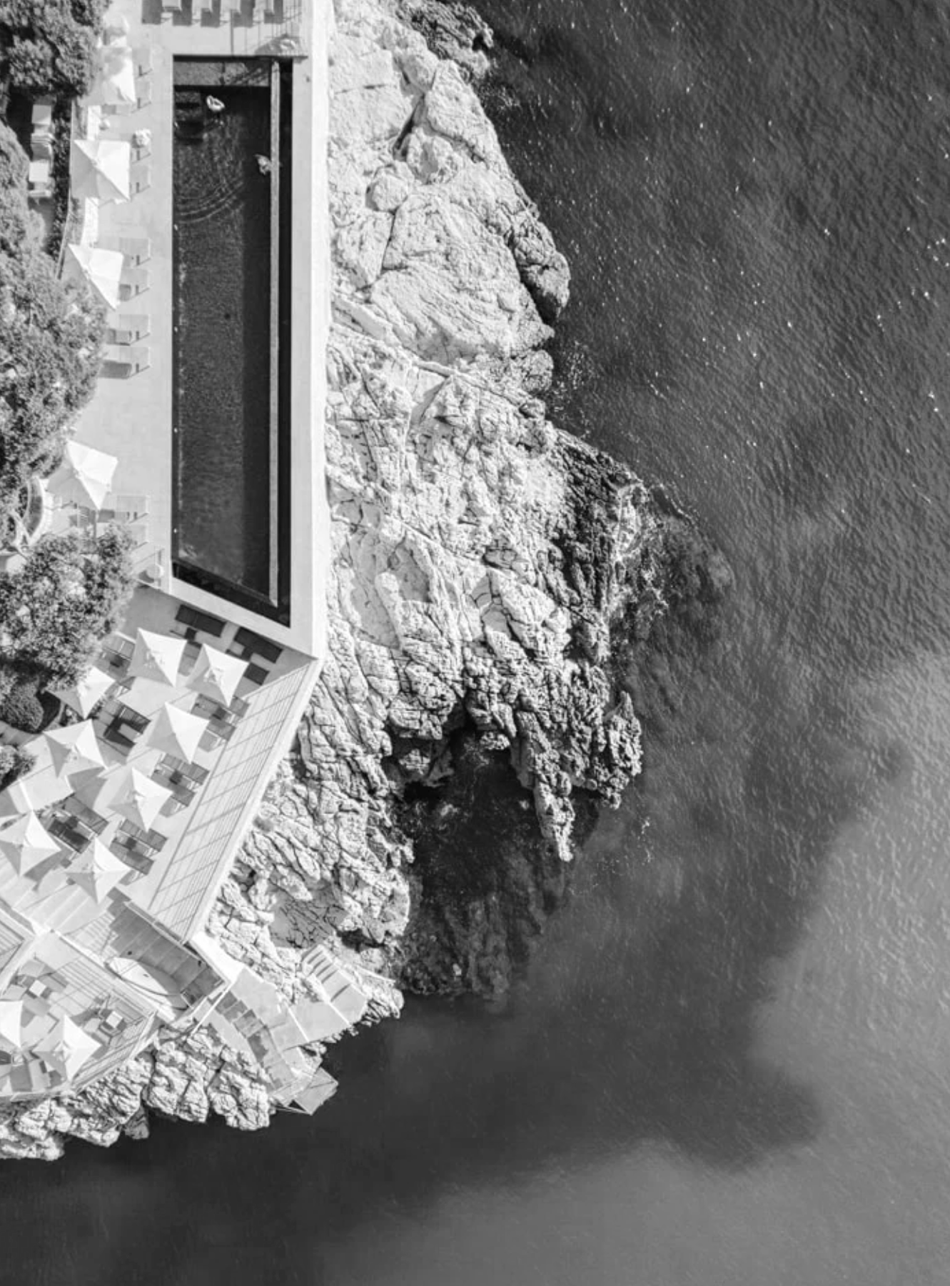 Aerial view of a seaside resort with a rectangular swimming pool, adjacent to rocky coastline and the ocean in black and white.