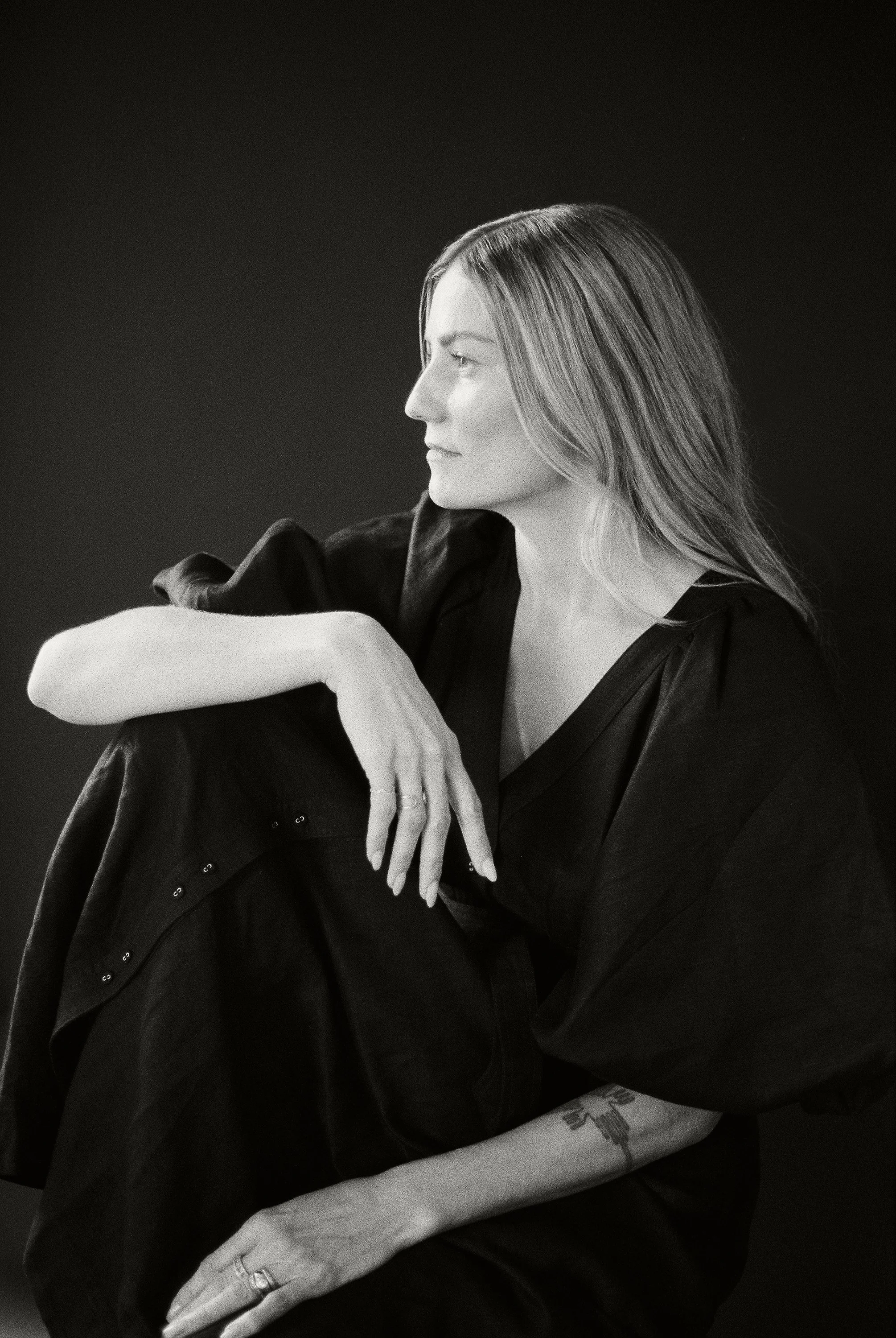 A black and white photo of a woman with long hair, wearing a dark jacket, sitting with her head turned to the side, resting her arm on her knee, and smiling slightly. She has a tattoo on her arm and wears rings on her fingers.