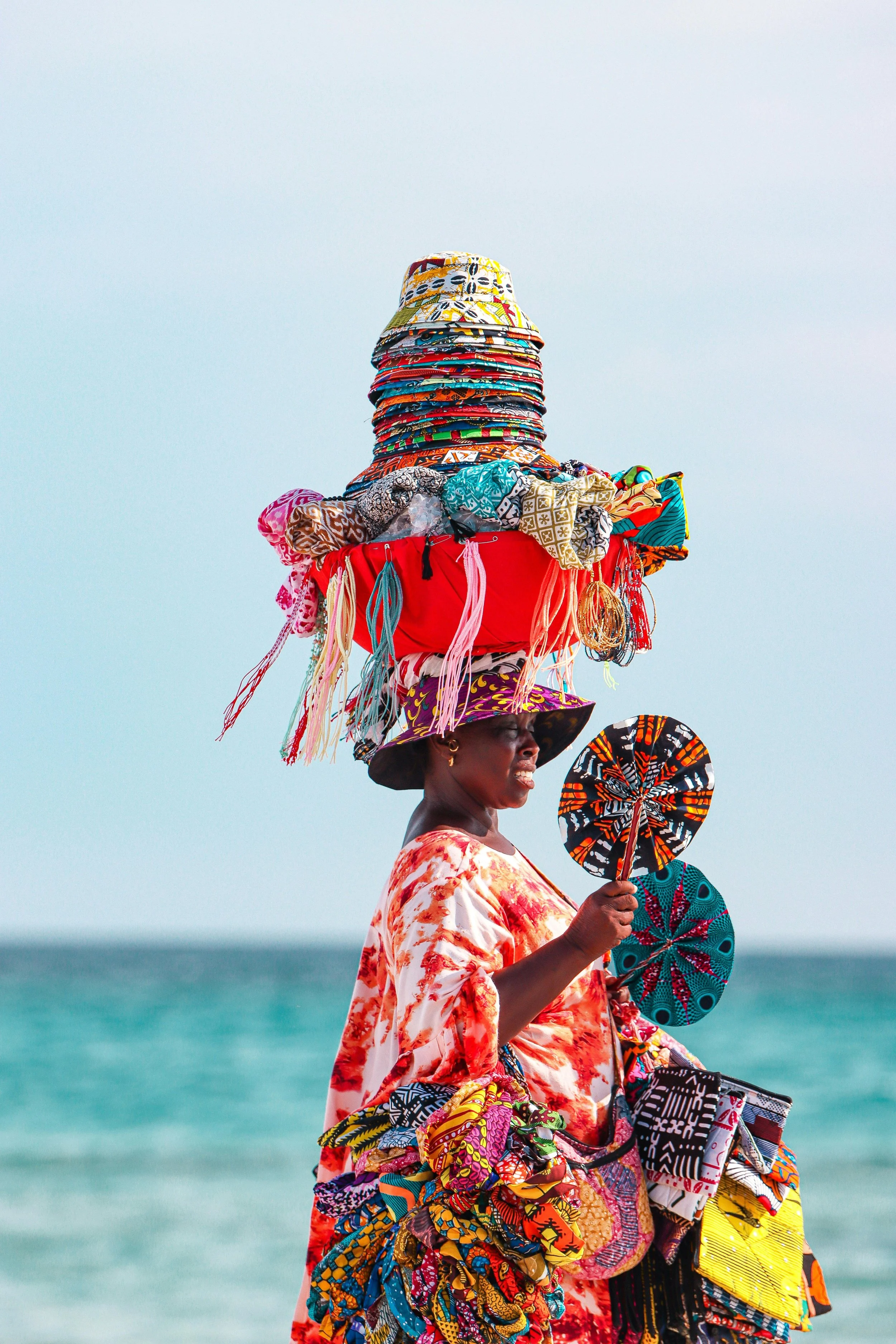 Femme portant un chapeau rempli de pagnes colorés en bord de mer.