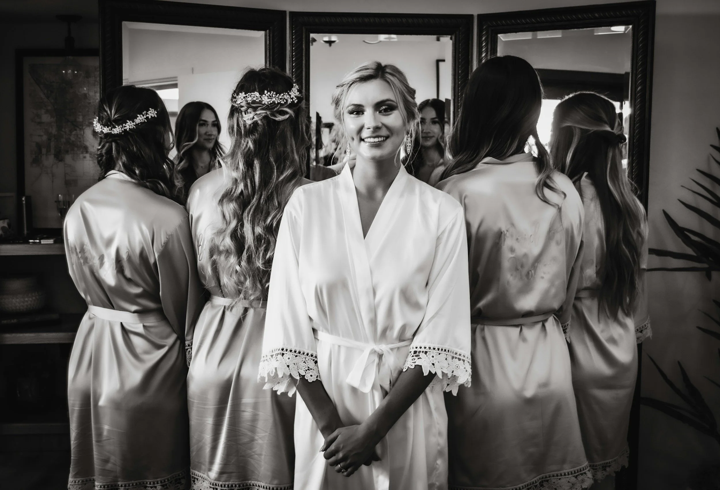 A woman dressed in a satin robe smiling at herself in a mirror surrounded by other women in similar robes, preparing for a special occasion.
