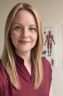 A woman with long blonde hair and blue eyes smiling, standing in front of a beige wall and a medical poster of the human anatomy.