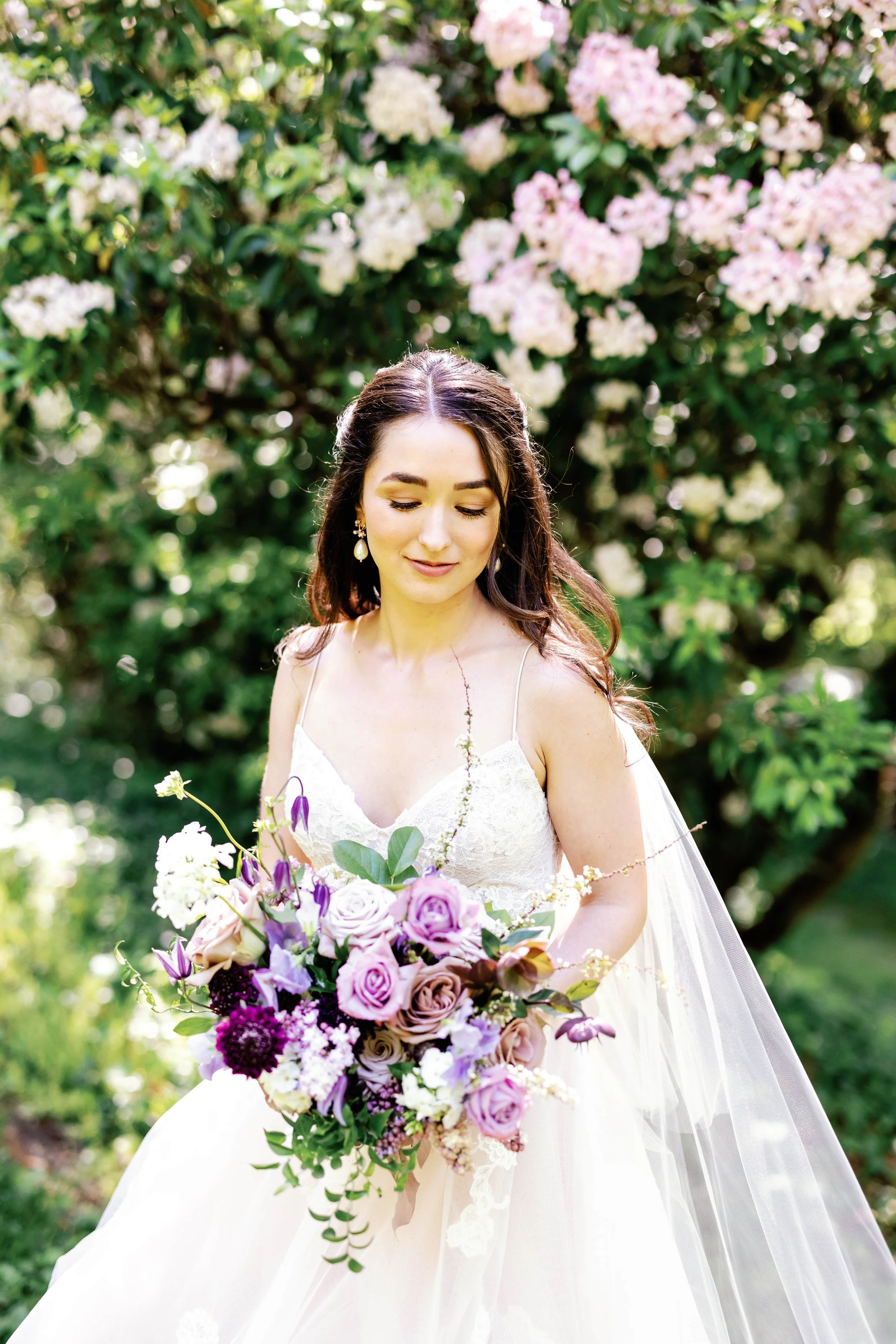 Bride in a white wedding dress holding a bouquet with pink and purple flowers, standing outdoors in front of a green floral arch on a grassy area, with trees and a house in the background.