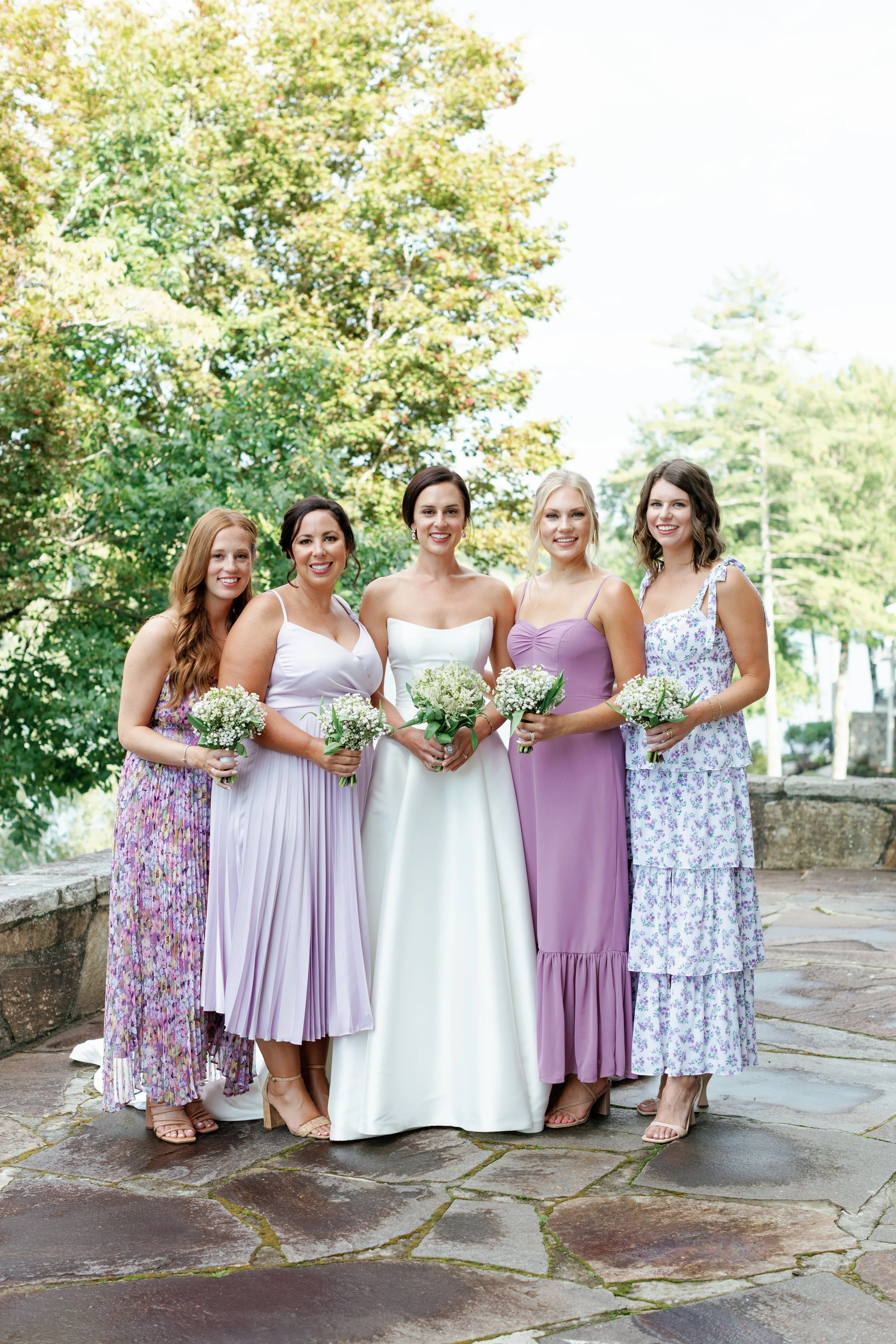 Group of  bridesmaids in colorful dresses standing outdoors, holding bouquets, with trees and a stone path in the background.