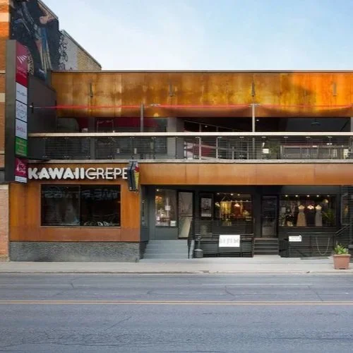 Exterior of Kawaii Crepe restaurant on a city street, two-story building with wooden and metal facade, glass doors, and large windows, with potted plant outside.