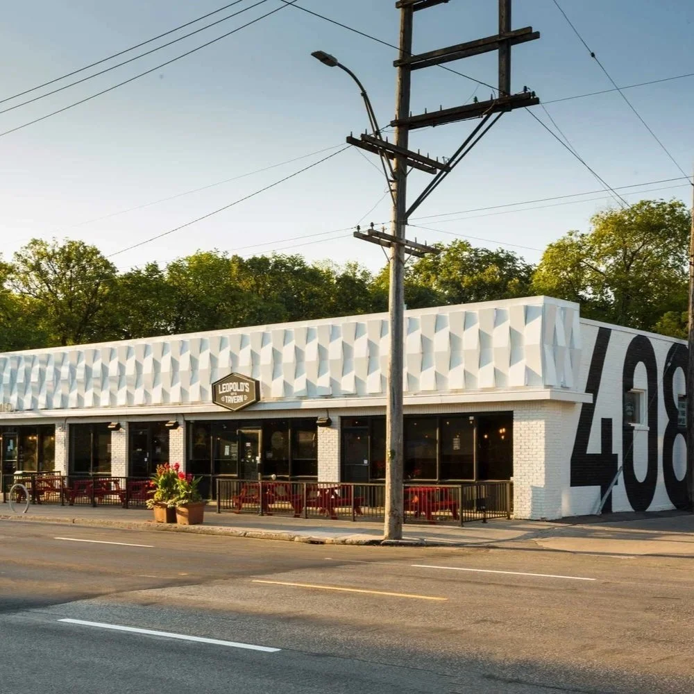 Restaurant named Leopold's with a modern white facade and large black numbers 408 on the side, outdoor seating with red chairs, sidewalk decor with potted plants, power lines overhead, and trees in the background.