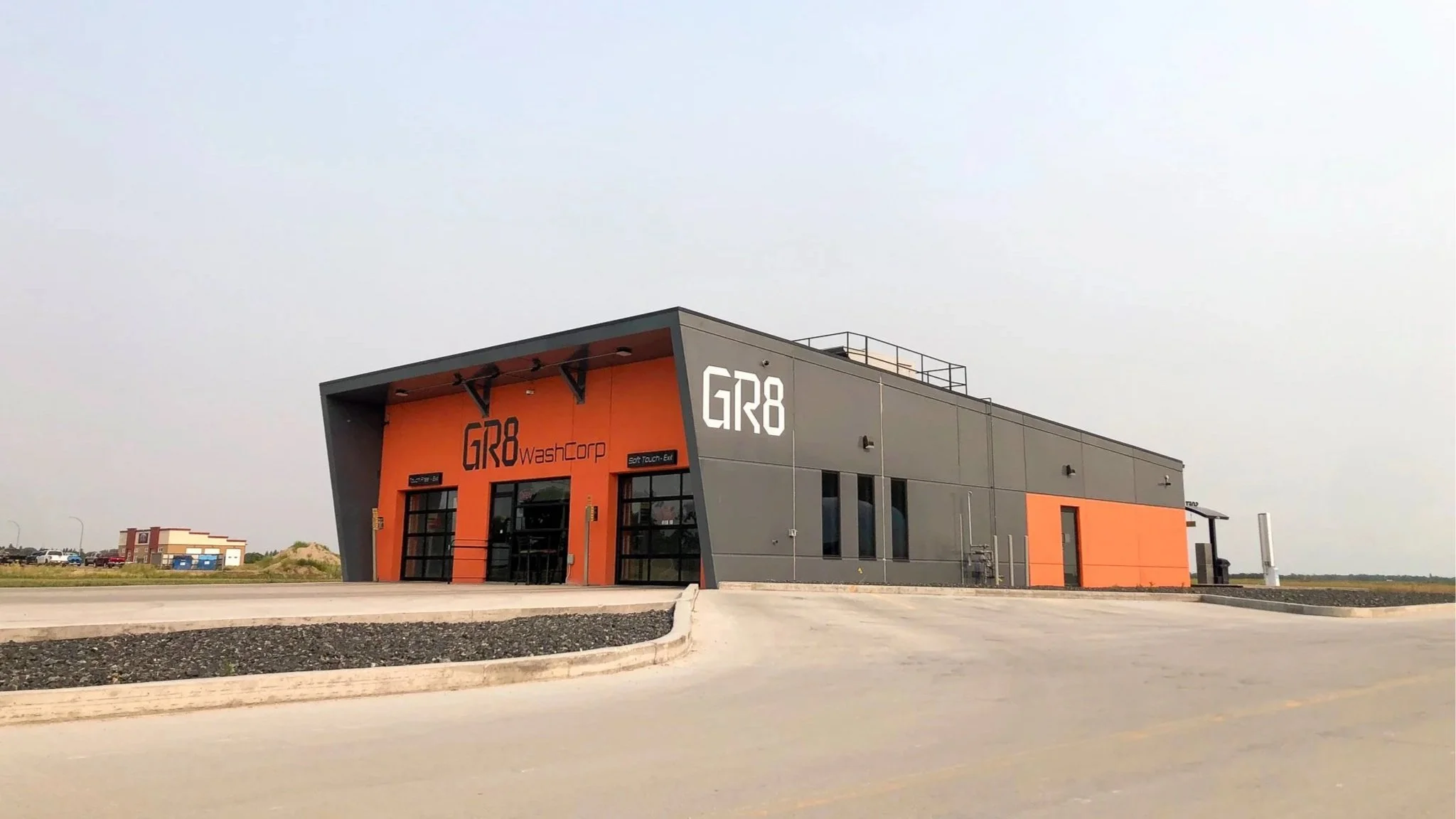 Modern industrial building with gray and orange exterior, labeled 'GR8' and 'WashCorp', situated in a spacious paved area under a cloudy sky.