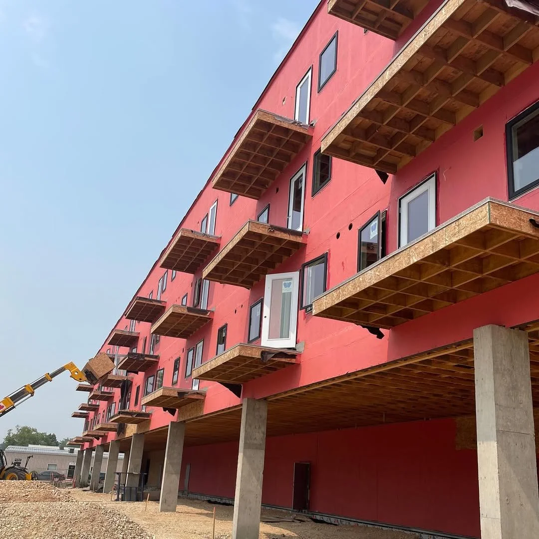 Under-construction multi-story building with pink exterior walls, small balconies, and vertical black window frames, supported by concrete pillars on the ground level, with construction equipment nearby.