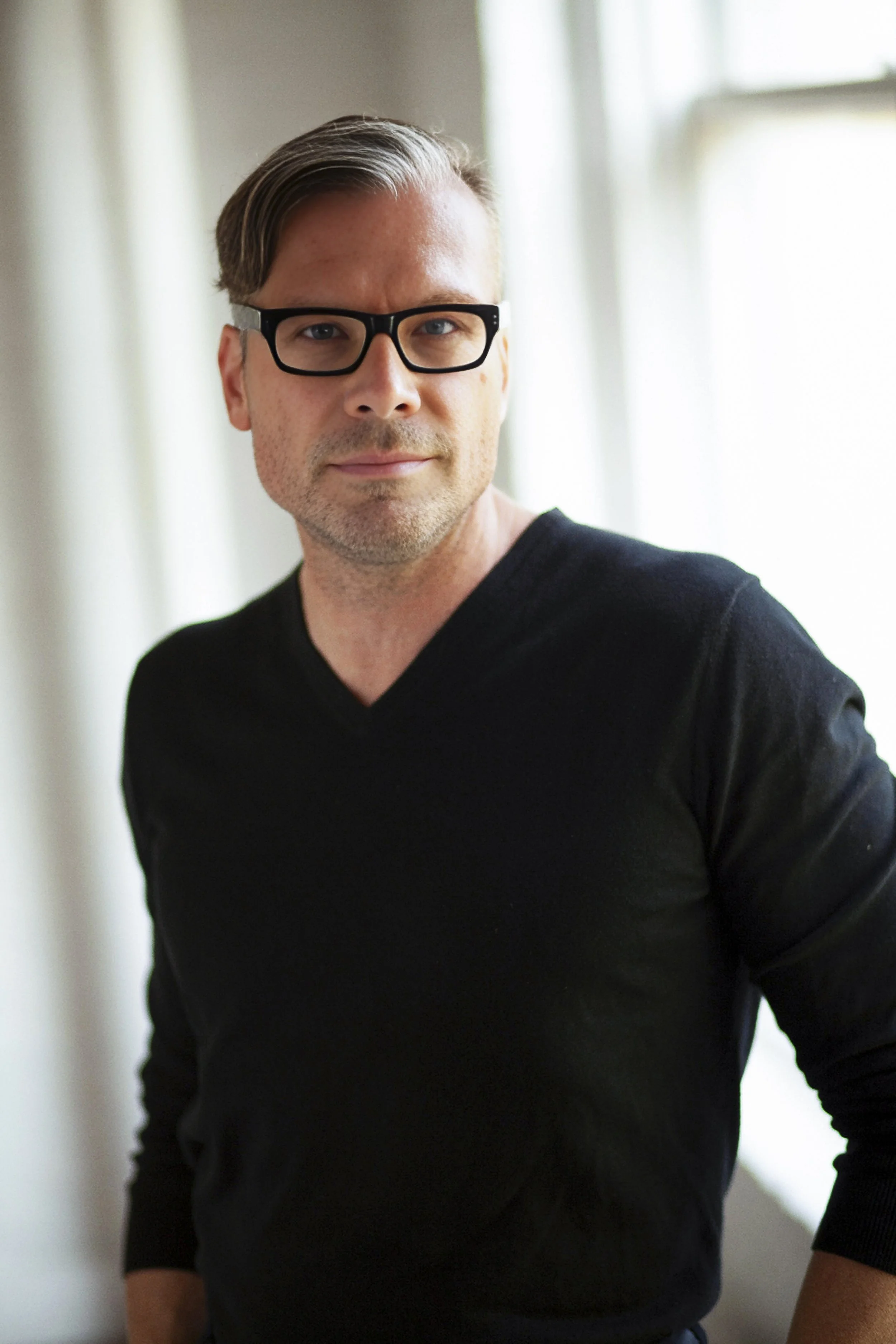 A man with glasses and light brown hair styled to the side, wearing a black V-neck shirt, standing indoors near a window with curtains, looking directly at the camera.