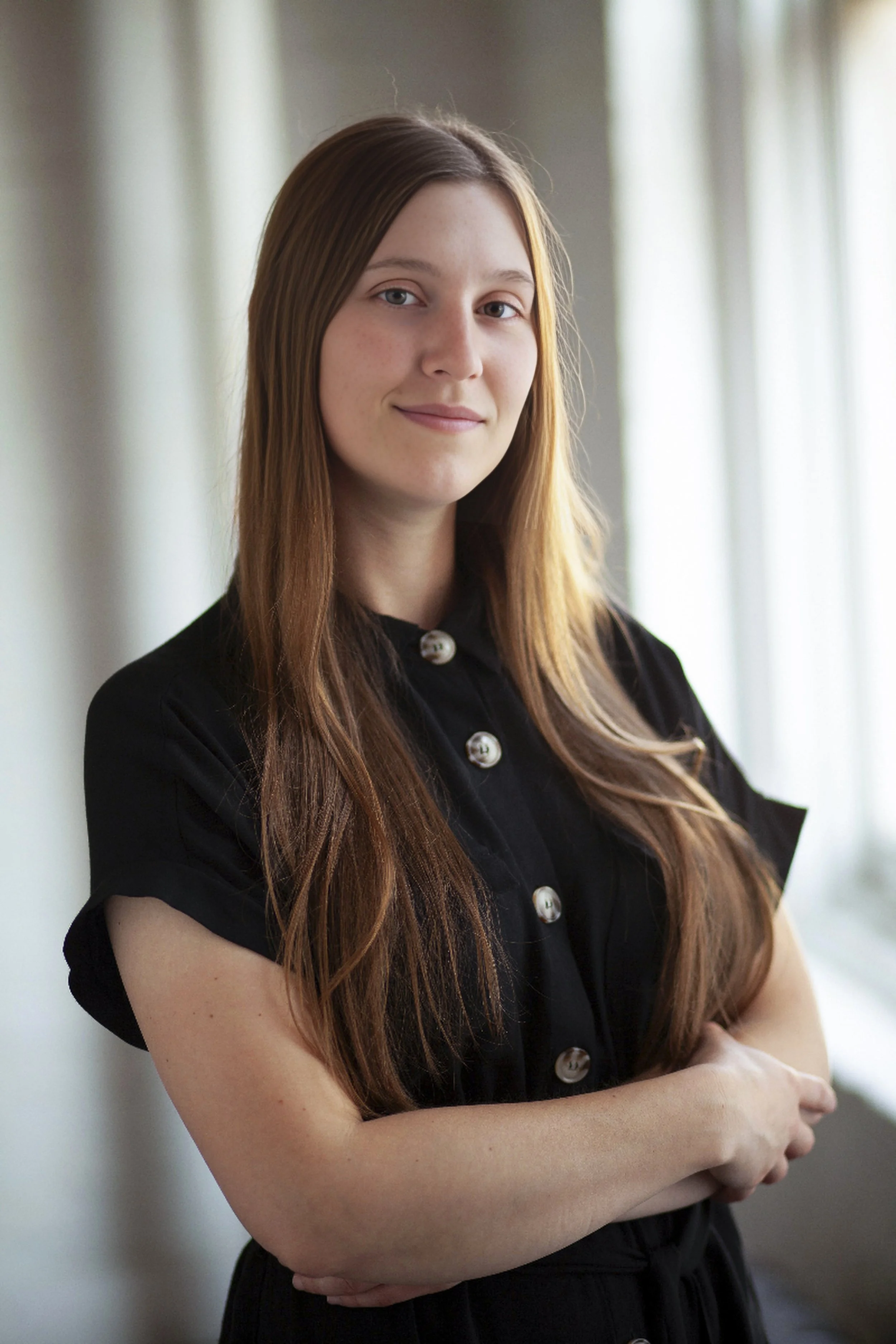 Young woman with long red hair wearing a black short-sleeved shirt with large buttons, standing indoors near a window with natural light.