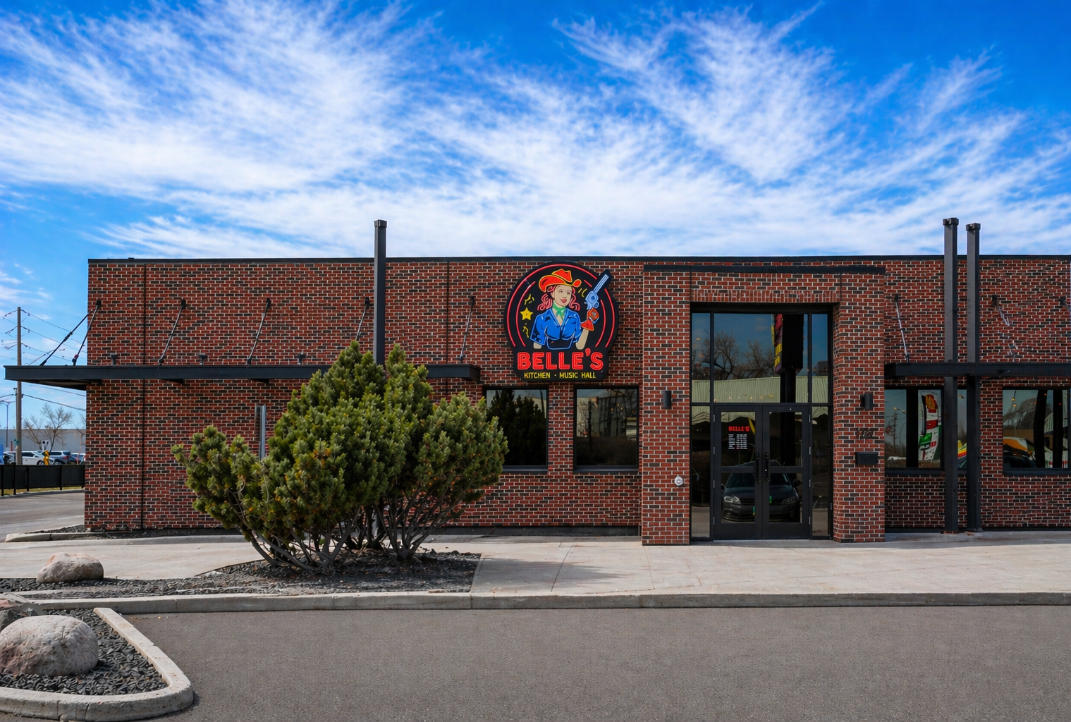 Brick building with a colorful neon sign of a woman in a cowboy hat holding a gun, labeled Belle's Kitchen and Music Hall, with a bright blue sky and some trees reflected in the windows.