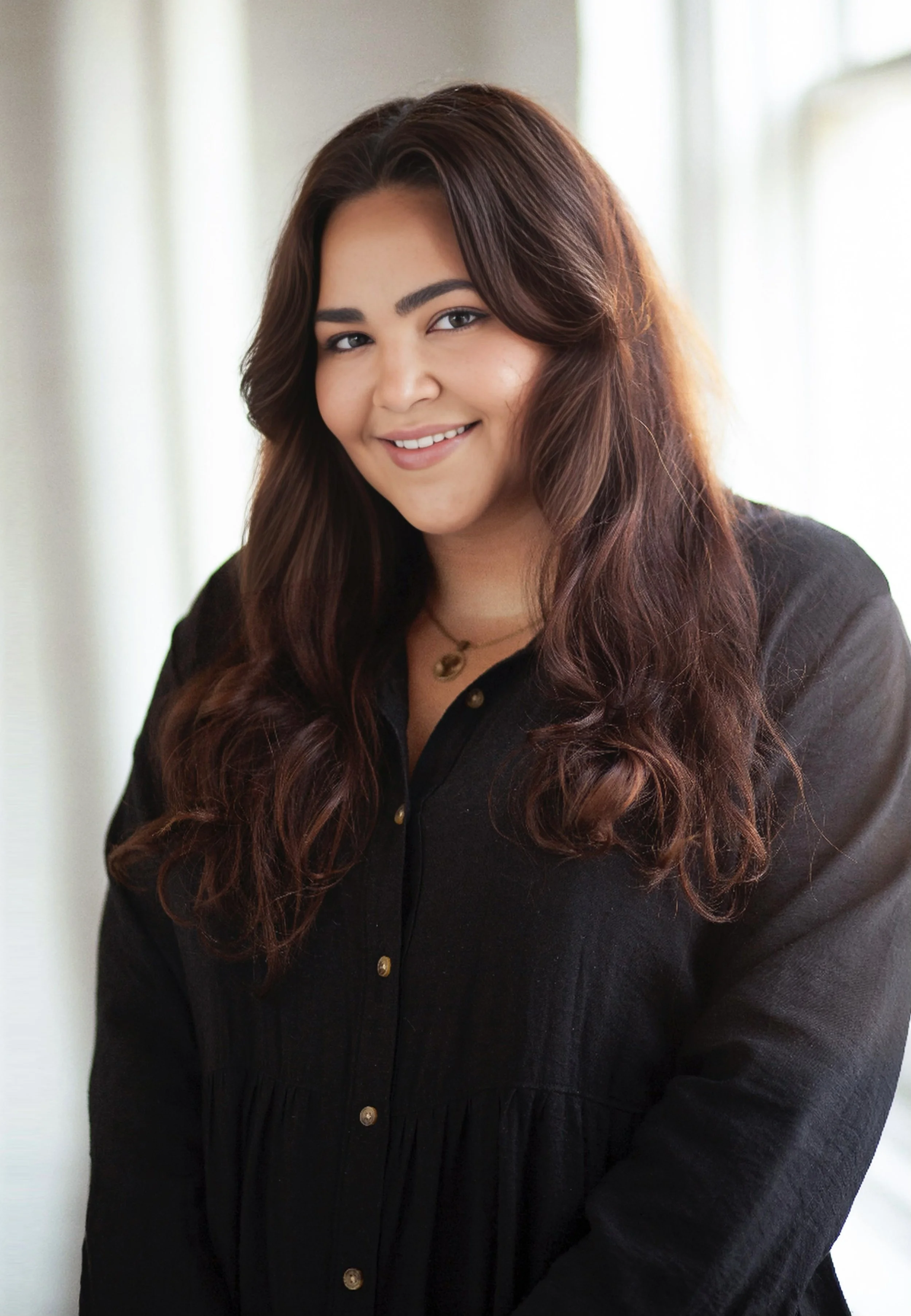 A young woman with long, wavy brown hair smiling while looking at the camera, wearing a black shirt with gold buttons and a necklace, standing near a window with light-colored curtains.