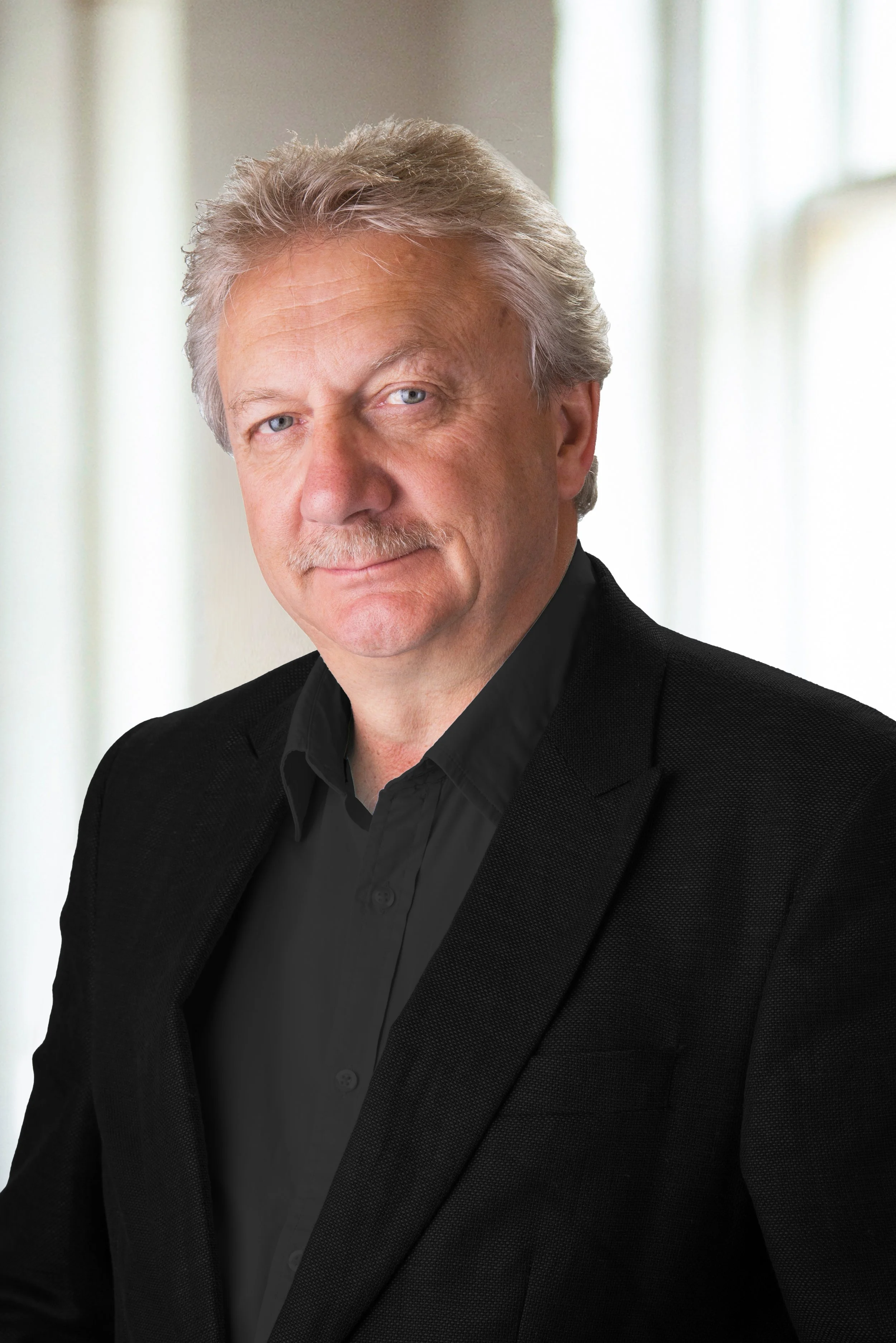 Photograph of an older man with gray hair and a mustache, dressed in a black blazer and shirt, standing in front of a softly lit background.