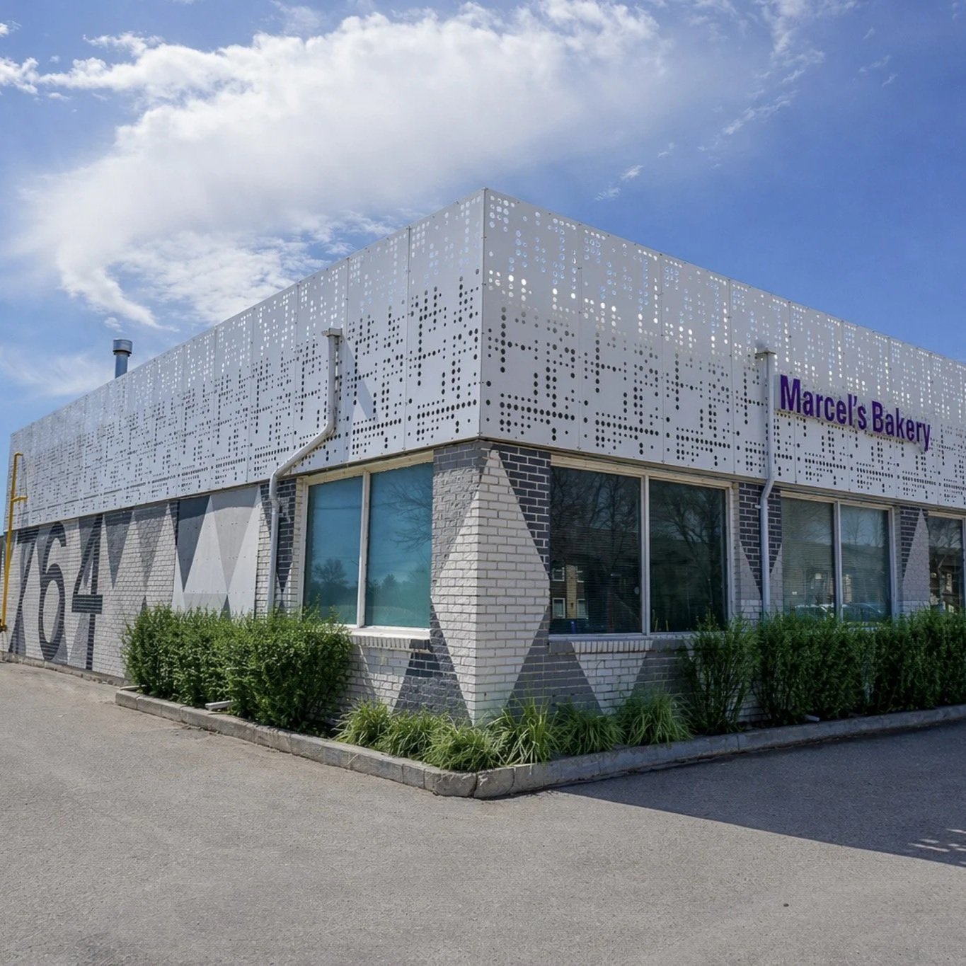 A modern commercial building with white perforated metal panels and brick walls, with the sign "Marcel's Bakery" on the front, situated on a bright day with a blue sky and sparse clouds. There are parked cars in front and small bushes along the building.