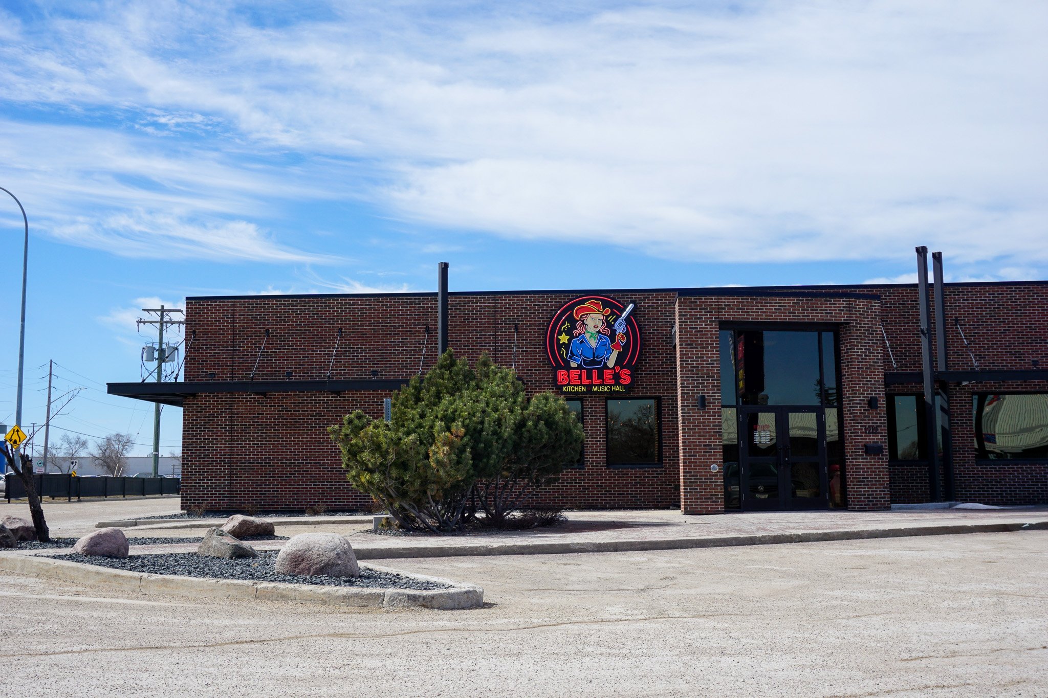 Brick building with a colorful neon sign of a woman in a cowboy hat holding a gun, labeled Belle's Kitchen and Music Hall, with a bright blue sky and some trees reflected in the windows.