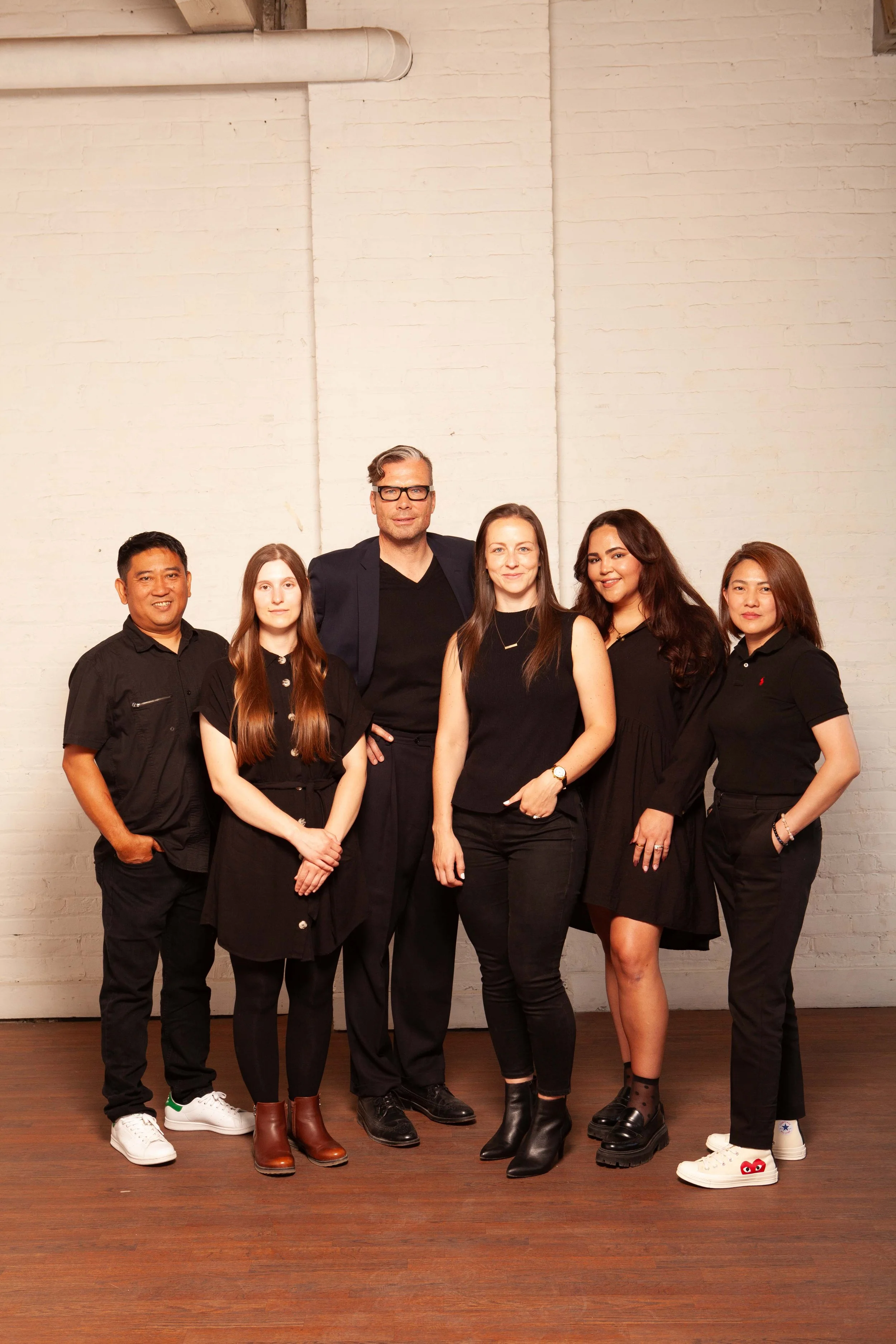 Group of six people standing together in front of a white brick wall, dressed in black and casual attire.