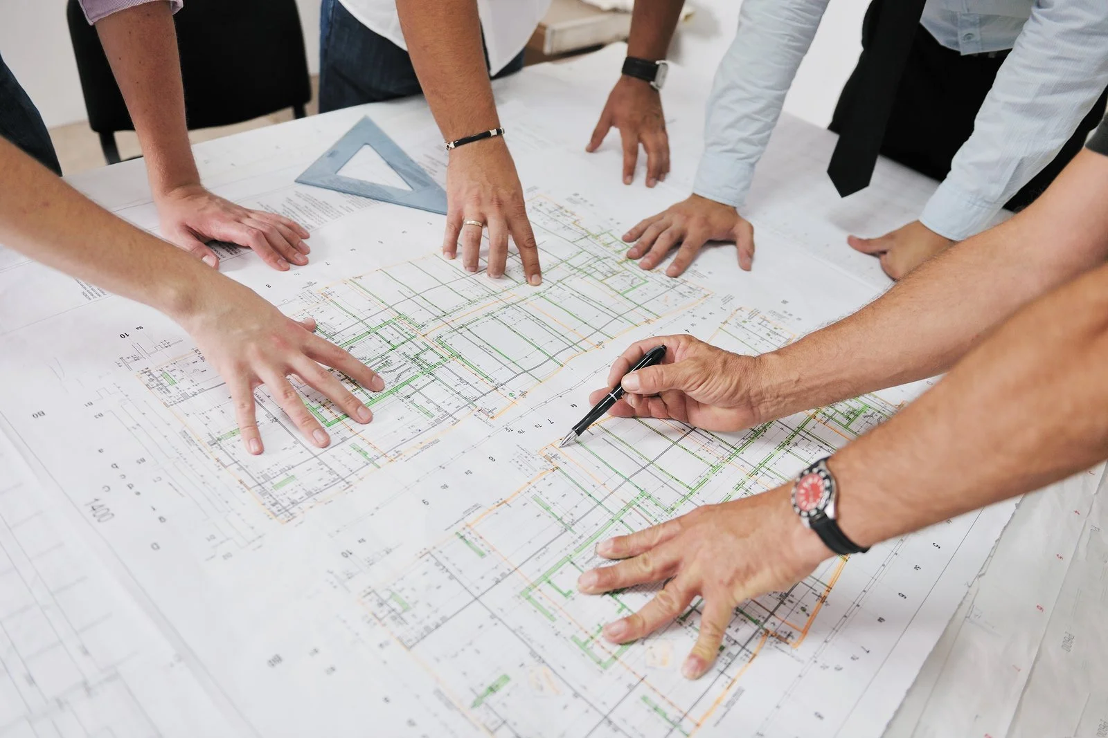 Several people are gathered around a large architectural blueprint on a table, examining and discussing the plans.