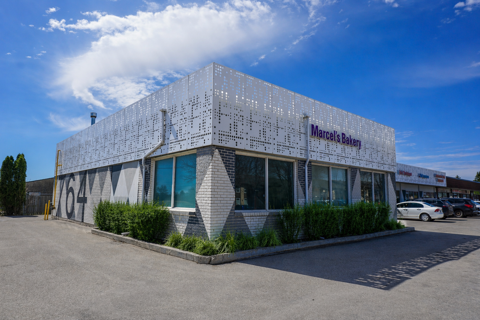 A modern commercial building with white perforated metal panels and brick walls, with the sign "Marcel's Bakery" on the front, situated on a bright day with a blue sky and sparse clouds. There are parked cars in front and small bushes along the building.