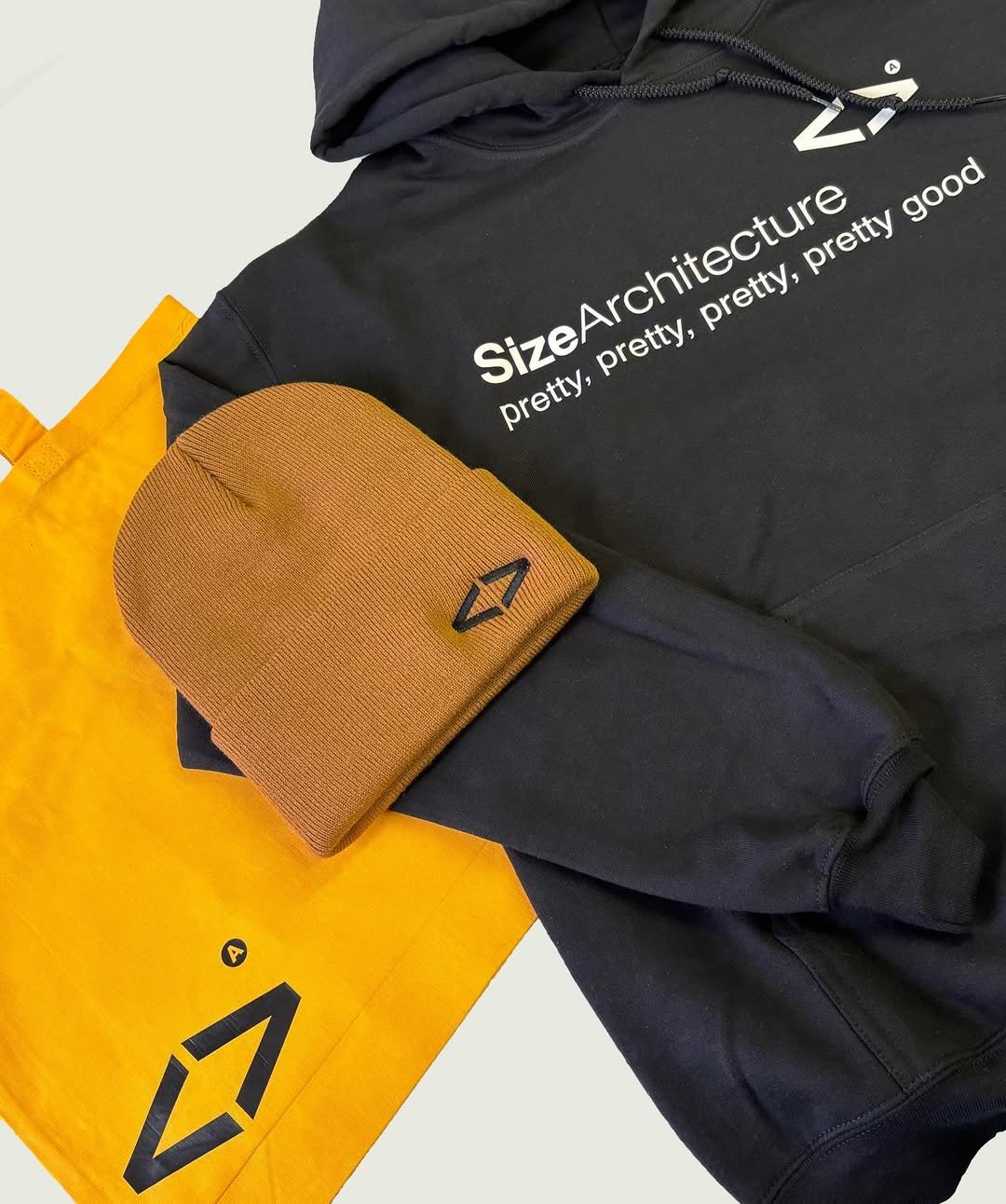 Black hoodie with white text that says 'SizeArchitecture Pretty, pretty, pretty, good', a brown beanie with black logo, and a yellow shopping bag with black graphics.