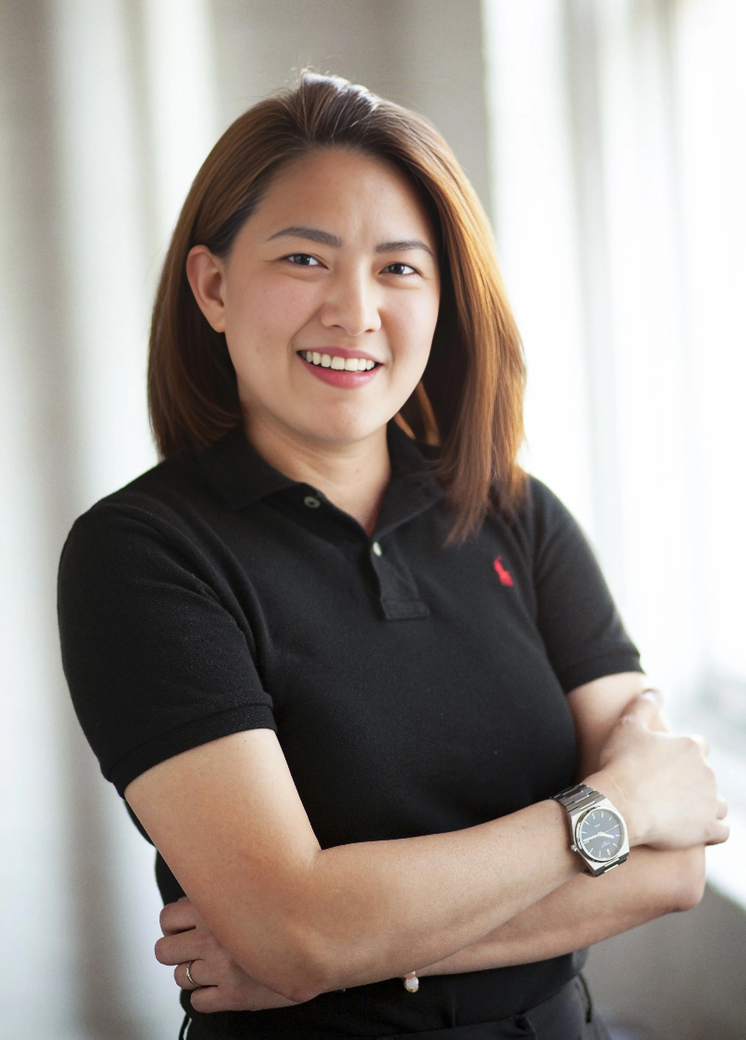 A woman with shoulder-length brown hair smiling, wearing a black polo shirt and a silver watch, standing indoors with light-colored walls and windows in the background.