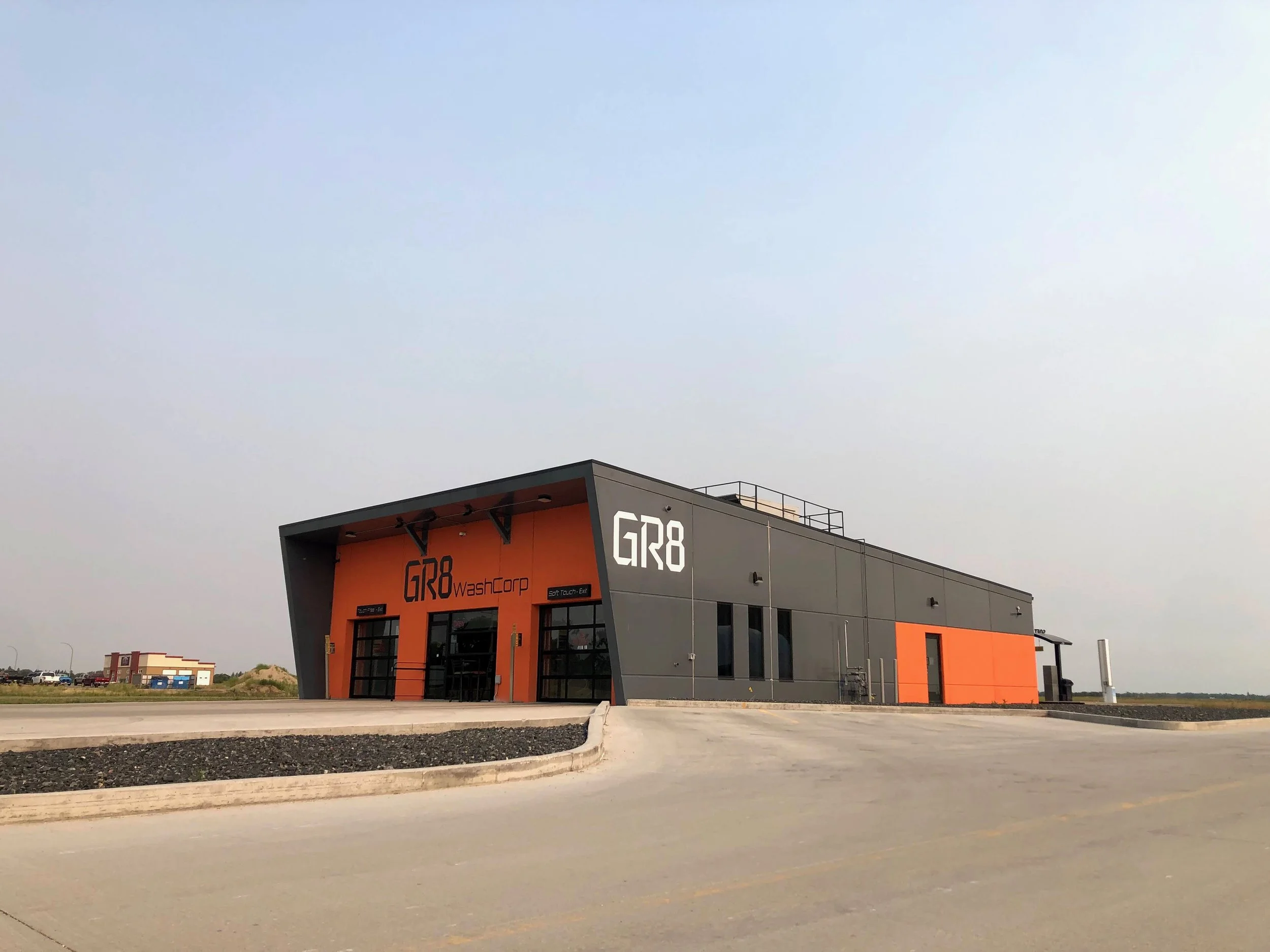 A large modern industrial building with the label 'GR8 Wash Corp' on the front, featuring a gray and orange exterior and a paved driveway leading up to the entrance.