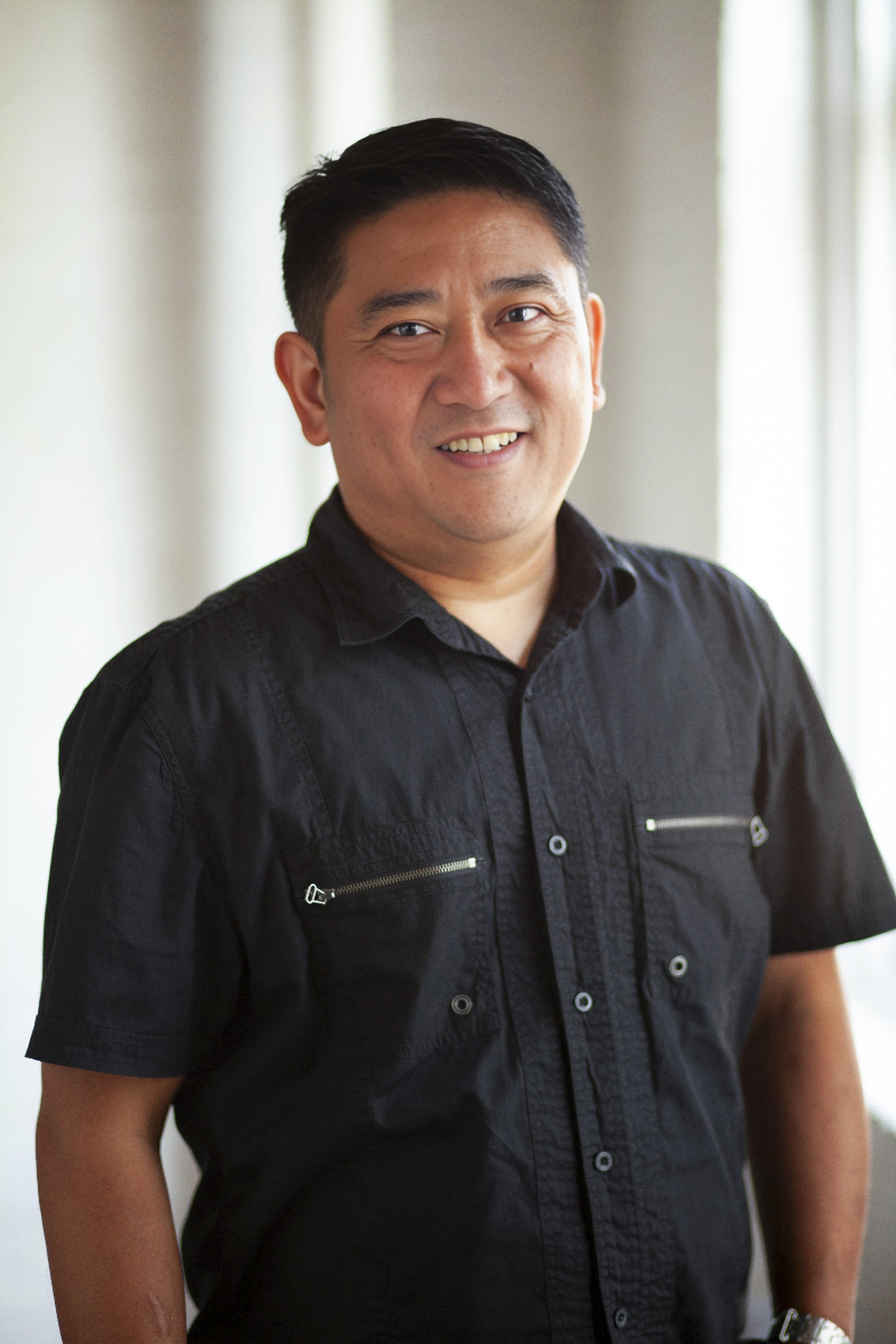 A smiling man with short black hair wearing a black short-sleeved button-up shirt standing indoors.