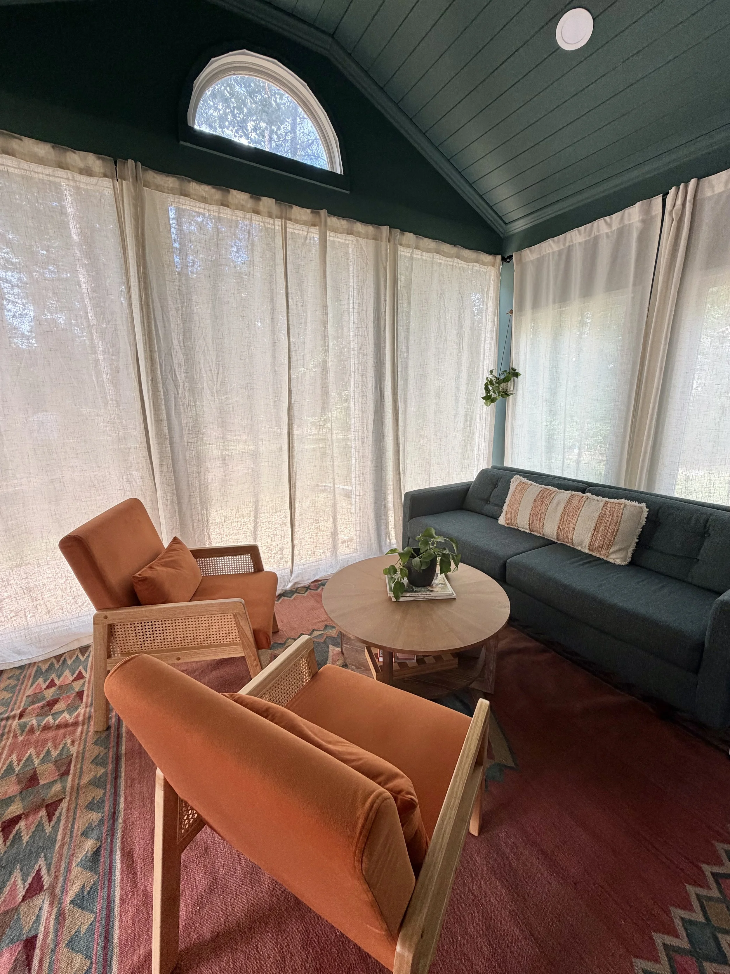 Cozy sunroom with dark green walls, sheer curtains, orange lounge chairs, a dark sectional sofa, round coffee table, and potted plants.