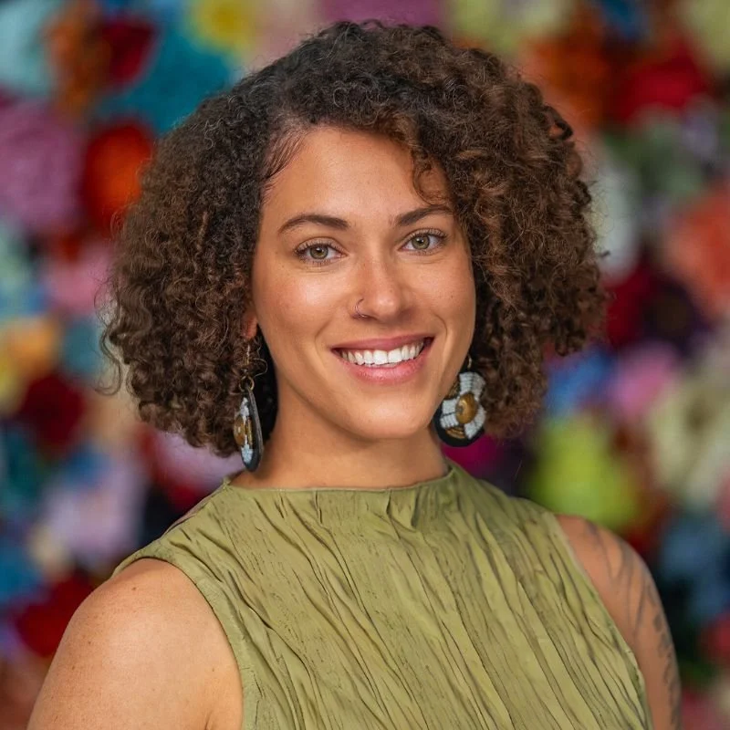 A young woman of color with curly brown hair, wearing a green sleeveless top and large earrings, smiling against a colorful blurred background.