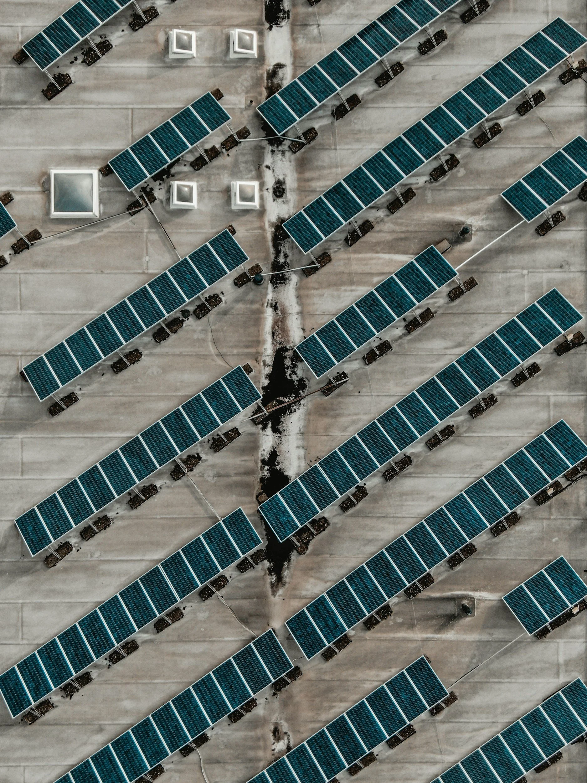 An aerial view of a solar panel installation on a flat roof, with rows of blue solar panels mounted on dark supports and some white ventilation units and patches of snow.