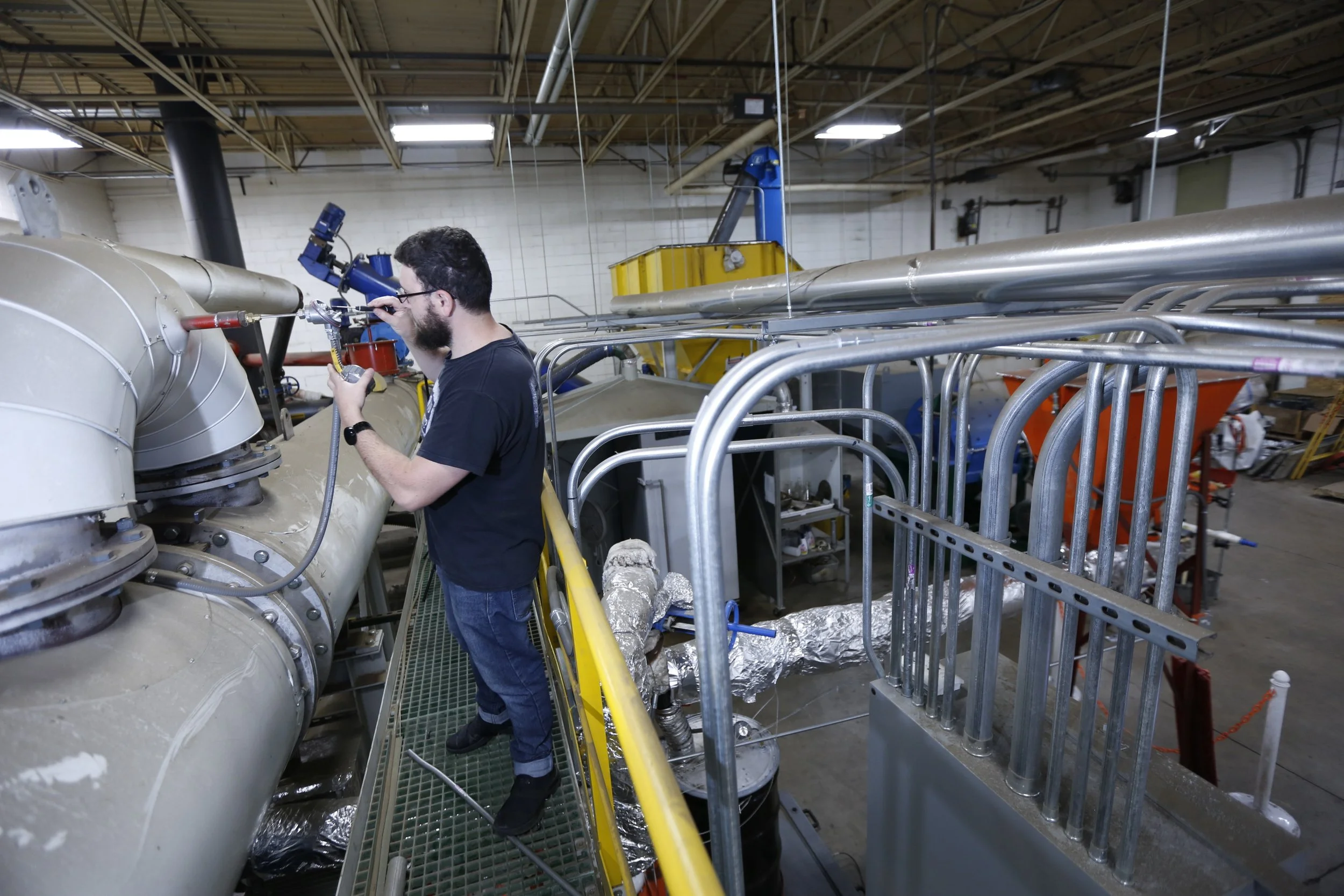 A technician with a beard and glasses working on a large industrial pipe in a factory or laboratory setting, surrounded by metal pipes, railings, and equipment.