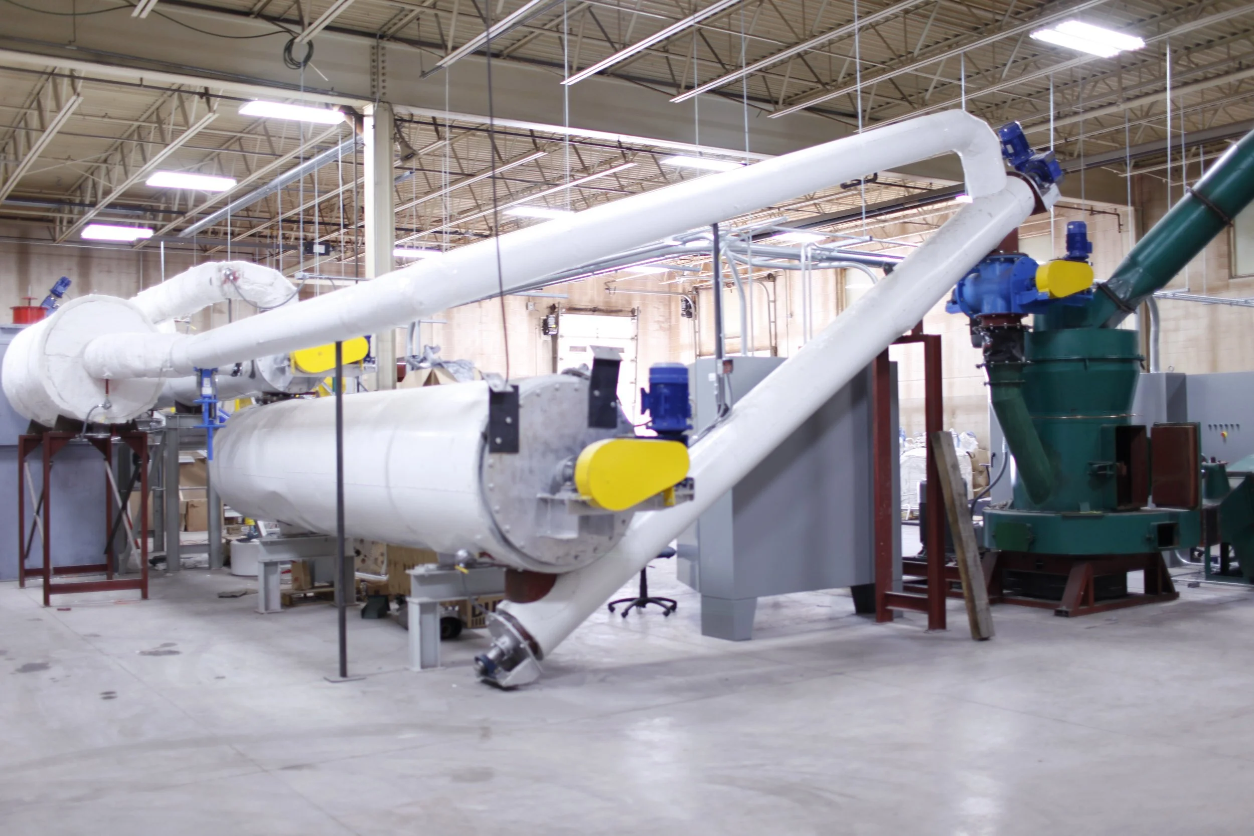 Industrial machinery with large white pipes, valves, and a green machine in a warehouse or factory setting.